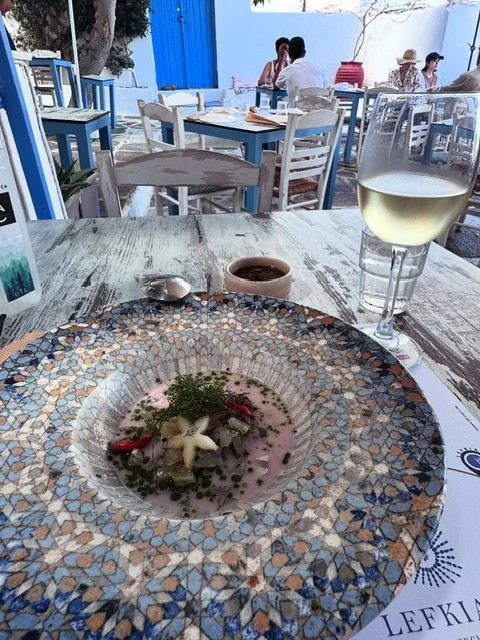 A dining table with a decorative plate, a glass of white wine, and a small bowl of sauce in an outdoor restaurant setting with other tables and people in the background.