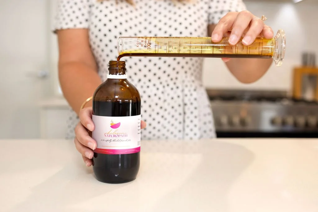Person pouring a yellow liquid from a graduated dropper into a brown glass bottle labeled "NATUROPATH" on a kitchen countertop.