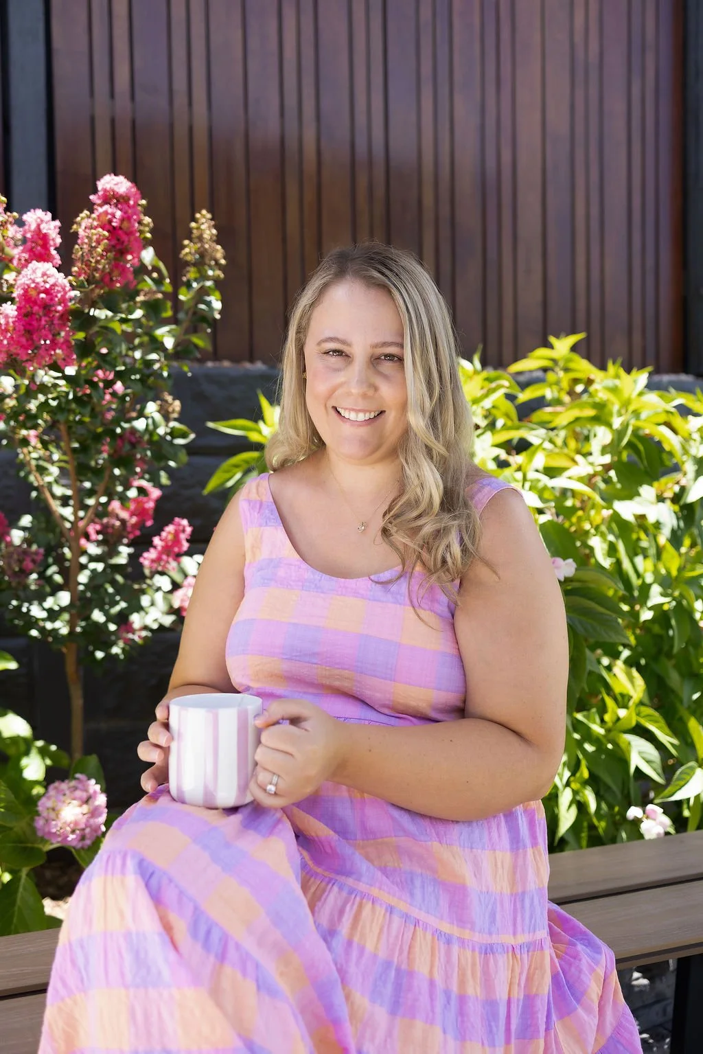A woman sitting outdoors on a wooden bench, smiling and holding a white and pink striped mug, surrounded by pink and green flowering plants, with a dark wooden fence in the background.