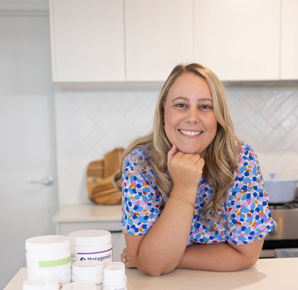 A woman smiling with her chin resting on her hand, sitting at a kitchen counter with supplement bottles in front of her.