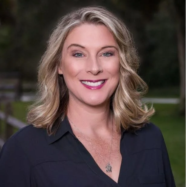A woman with shoulder-length blonde hair smiling outdoors, wearing a black shirt and a necklace.