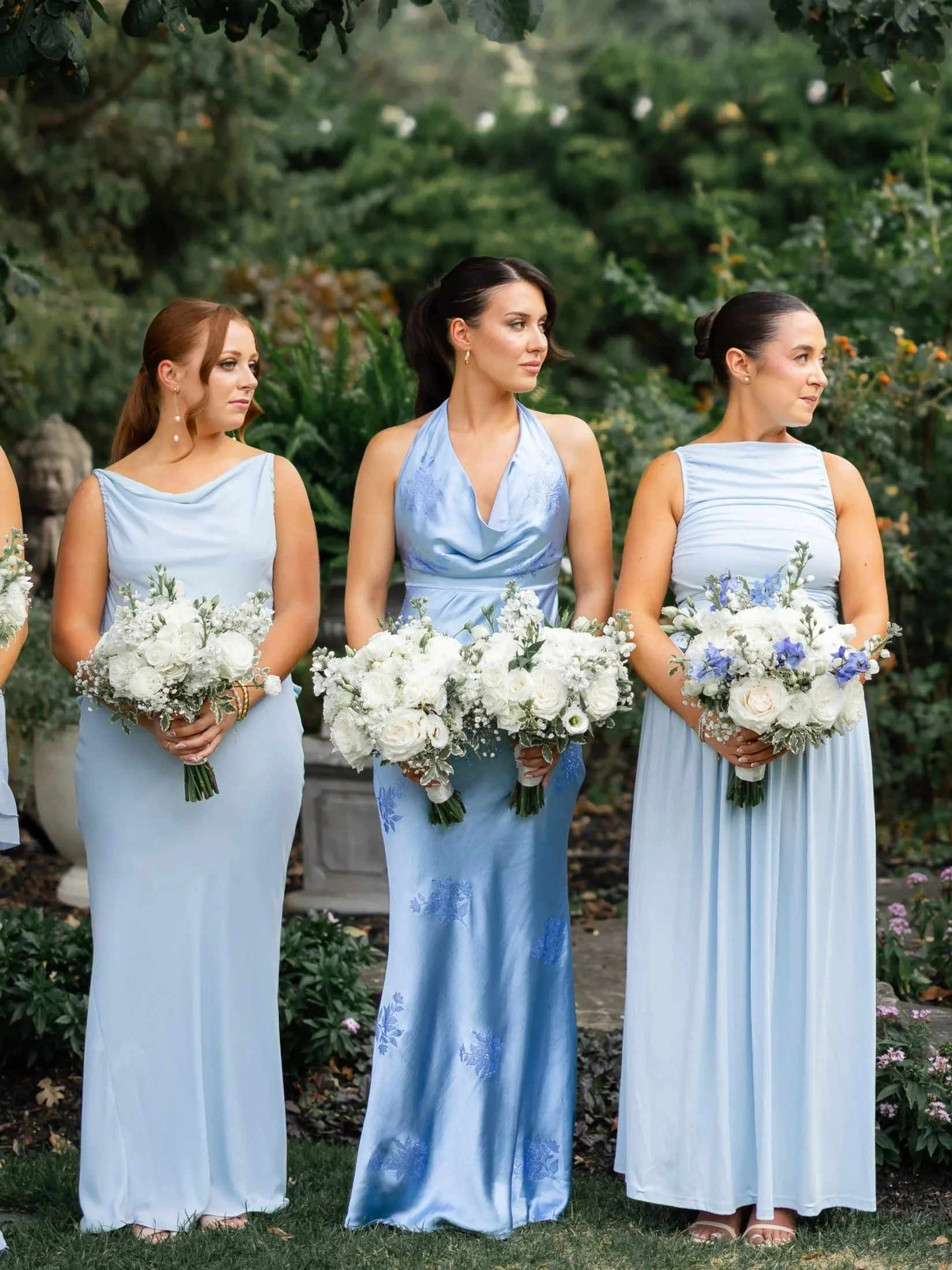 bridesmaids in light blue dresses standing outdoors under a large tree, holding white floral bouquets, preparing for a wedding or formal event. toronto wedding vendors, luxury bridal makeup artist, natural soft glam.
