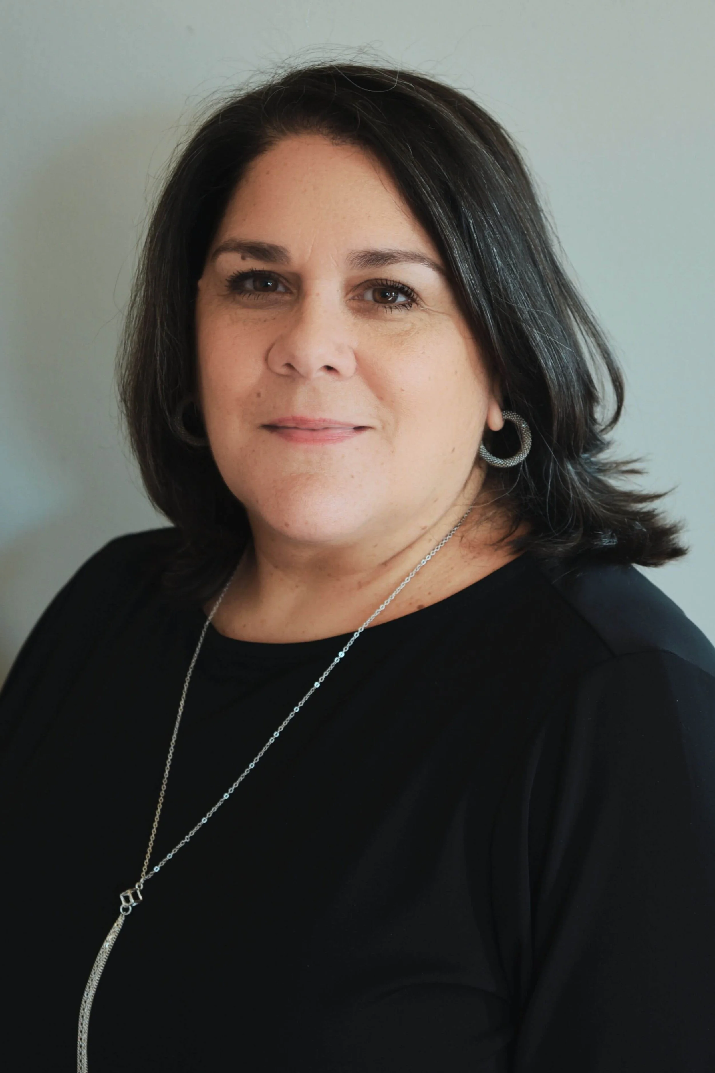 A woman with dark hair, wearing a black top, silver earrings, and a silver necklace, standing against a plain background.