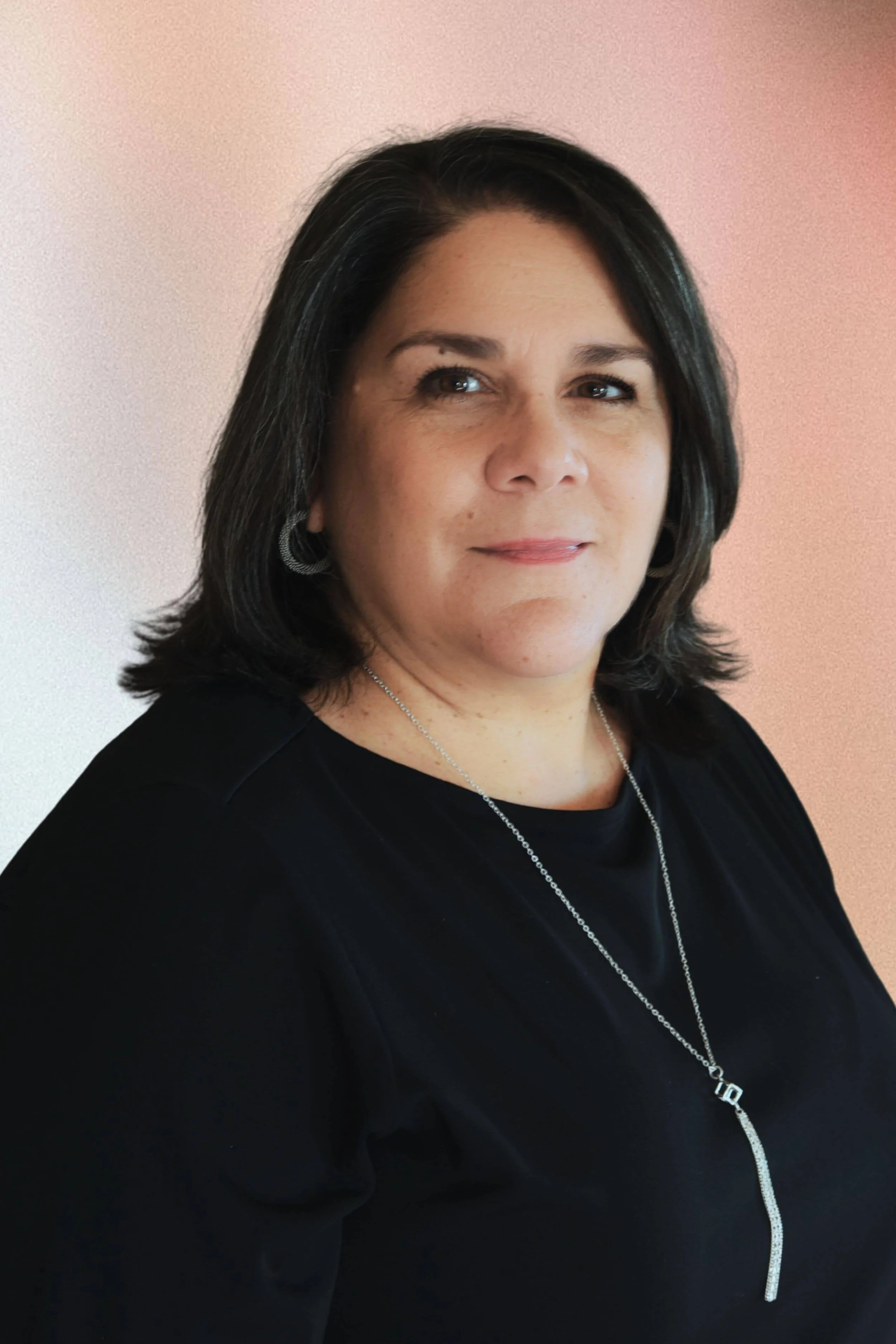 A woman with shoulder-length dark hair, wearing a black top, silver earrings, and a silver necklace with a pendant, standing against a pastel wall.
