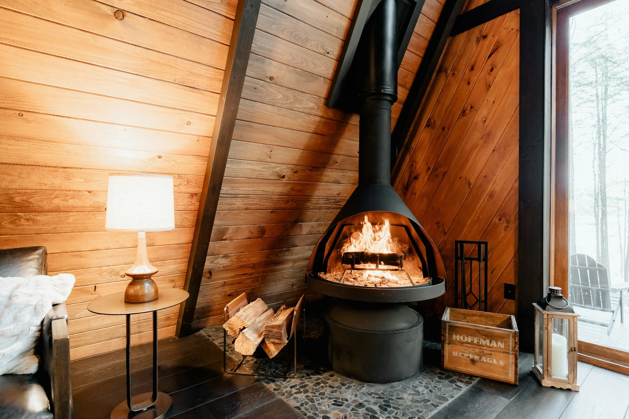 Interior of a cozy wooden room with a modern black fireplace burning fire, a side table with a lamp, a crate labeled 'Hoffman Beverages', a lantern with a candle, and a large window revealing an outdoor deck and trees.