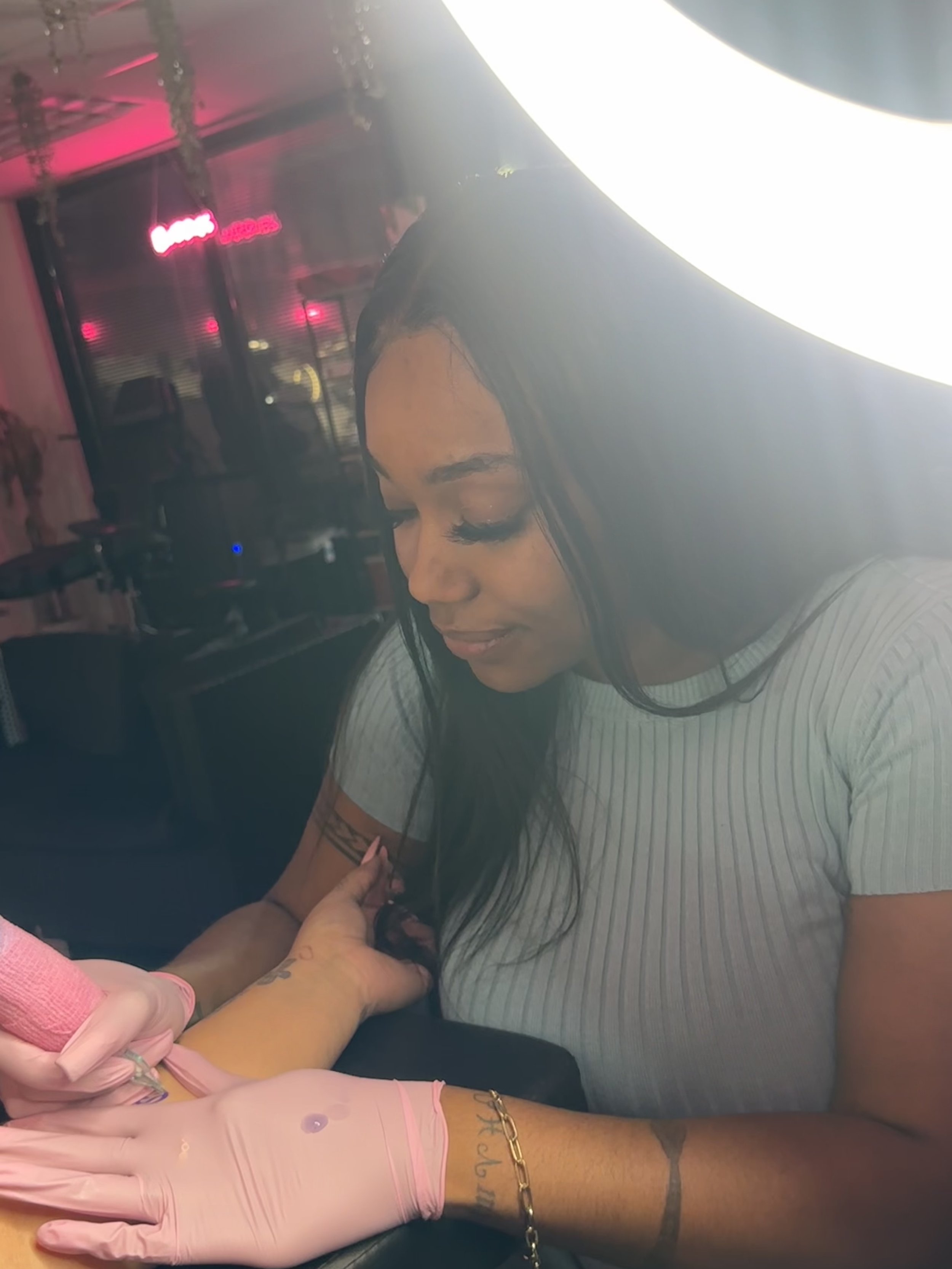 A woman with long black hair getting a tattoo on her arm at a tattoo parlor with pink neon lights in the background.