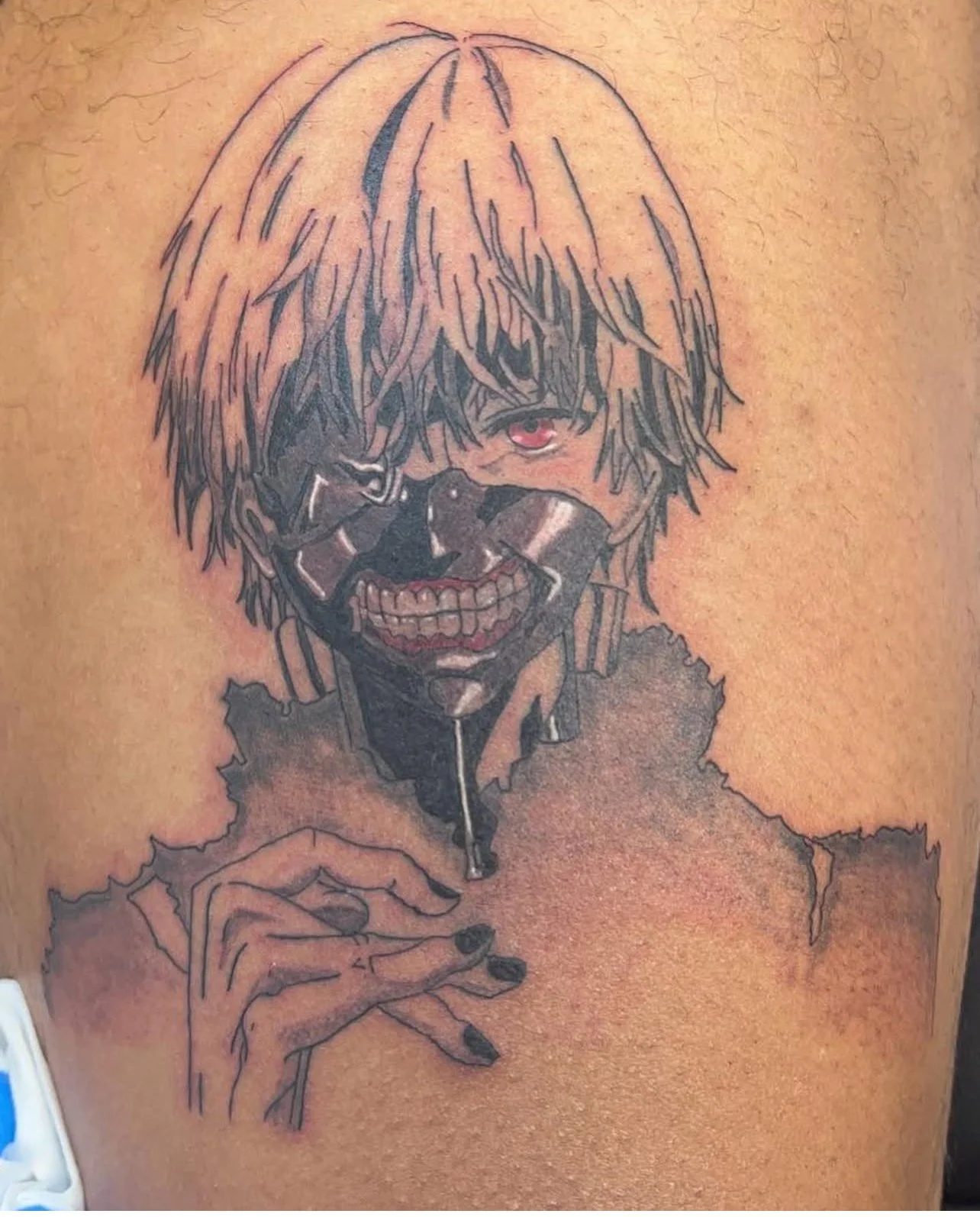 Tattoo of a person with blond hair wearing a metallic mask covering the lower face, one eye visible with red iris, holding a cigarette between fingers. Ken kenaki from Tokyo Ghoul.