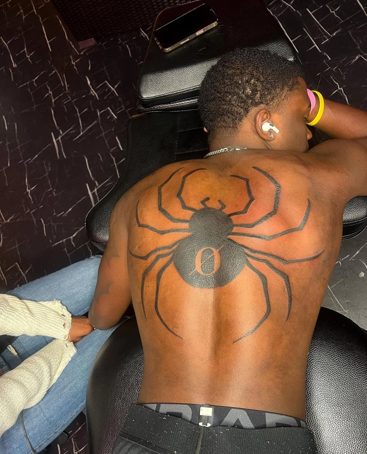 A person with short hair and earbuds lying face down on a black leather chair, with a large spider tattoo on their back featuring the number 0 in the center from HunterxHunter