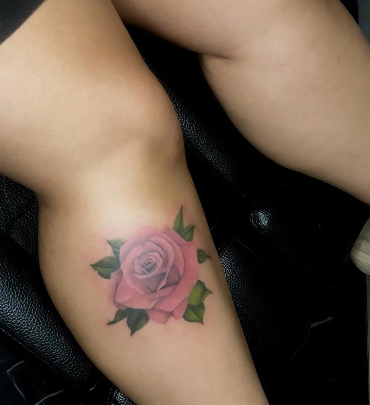 A tattoo of a pink rose with green leaves on a person's upper thigh.