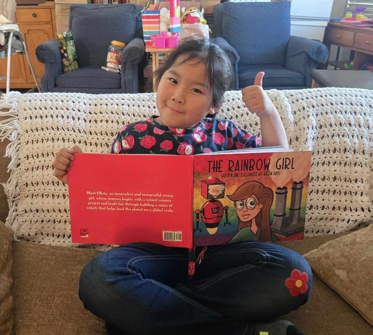 A young girl sitting on a couch, holding an illustrated children's book titled 'The Rainbow Girl.'