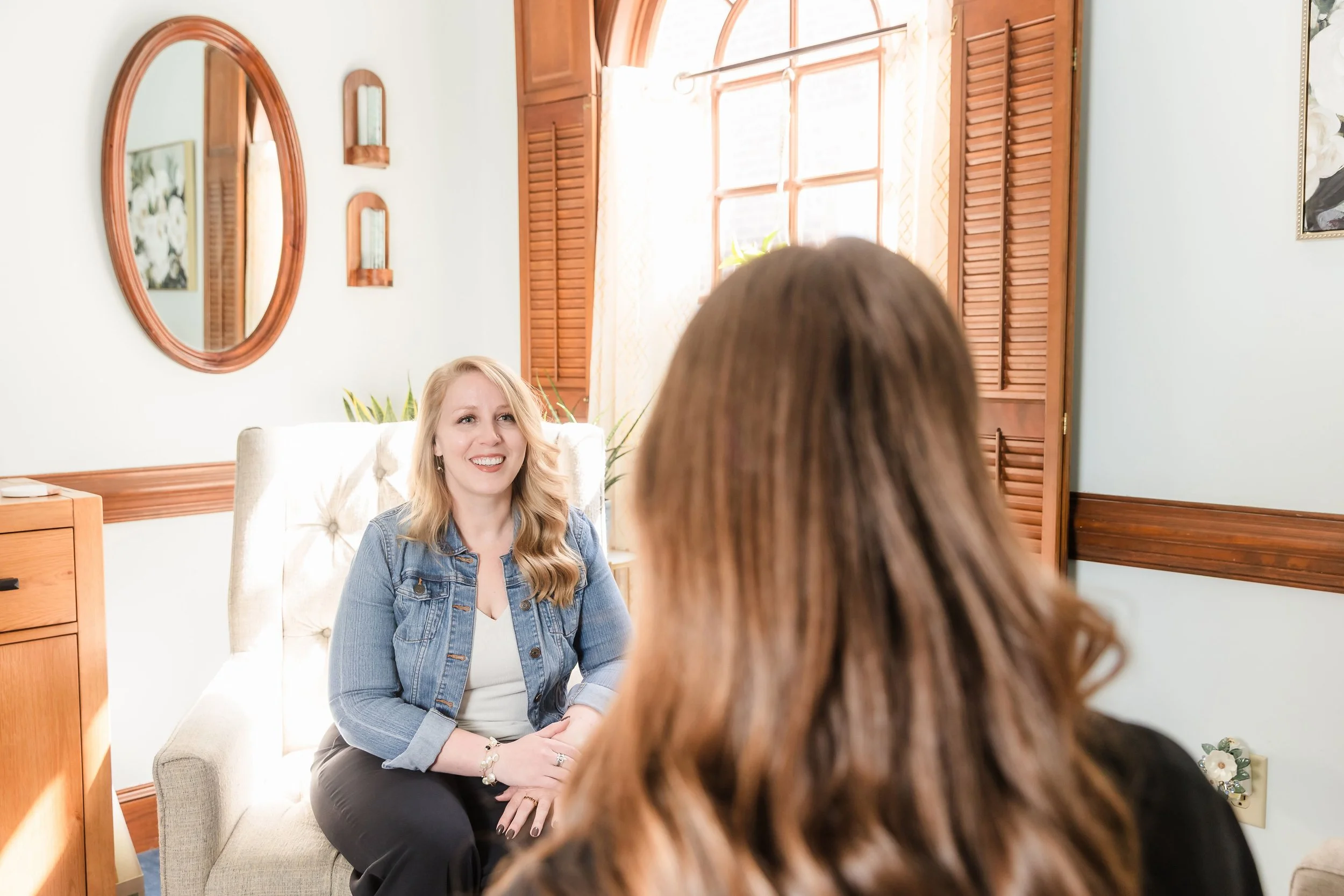 Two women having a conversation in a bright, cozy room with wooden accents and natural light. One woman with blonde, wavy hair wearing a denim jacket sits on a beige armchair, smiling. The other woman has brown hair and is facing away from the camera.