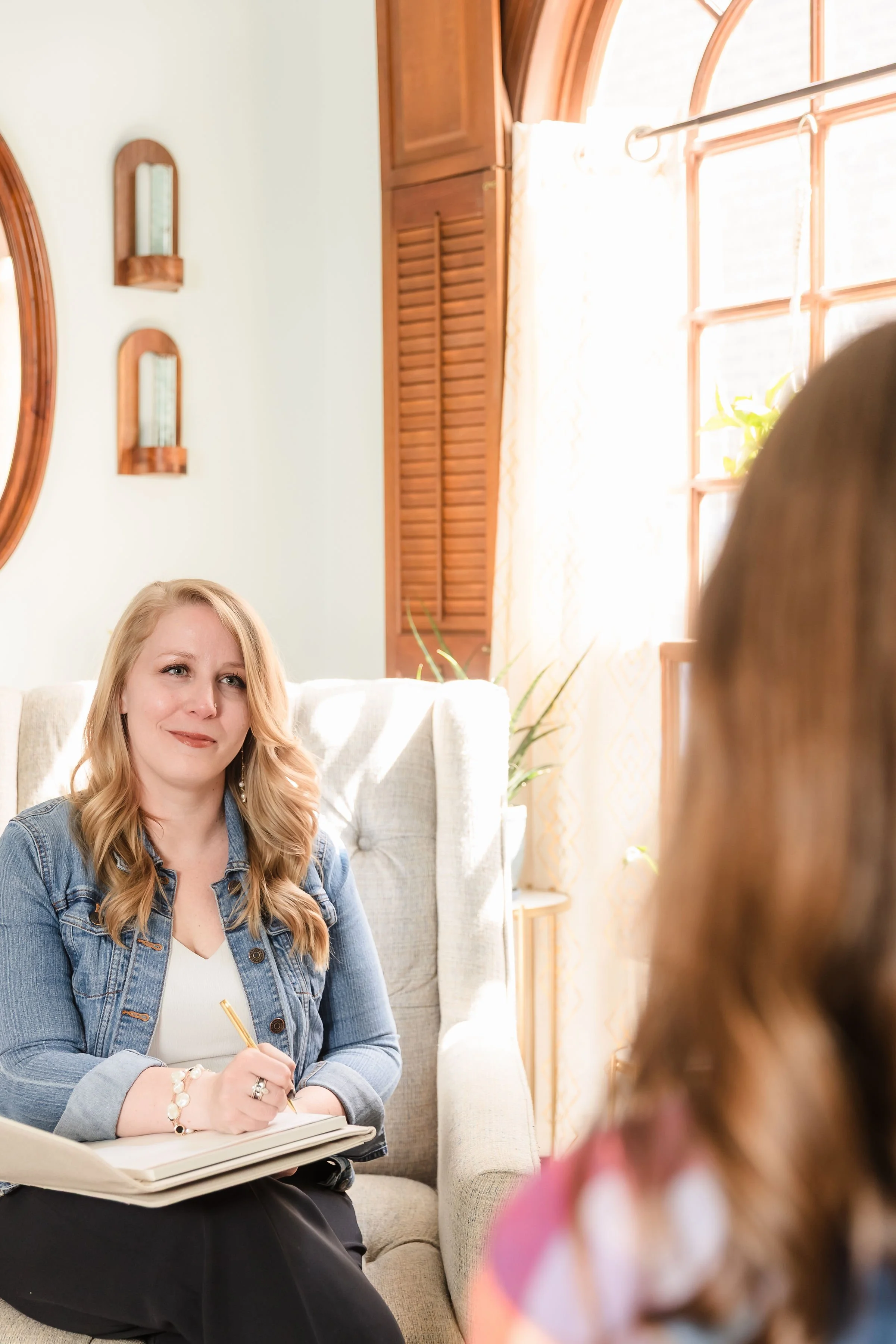 Dr. Jamie L. Chachere, a therapist with blonde wavy hair sitting on a beige armchair, holding a notepad and a pen, engaging in a conversation in a sunlit room with wooden window frames and decor.