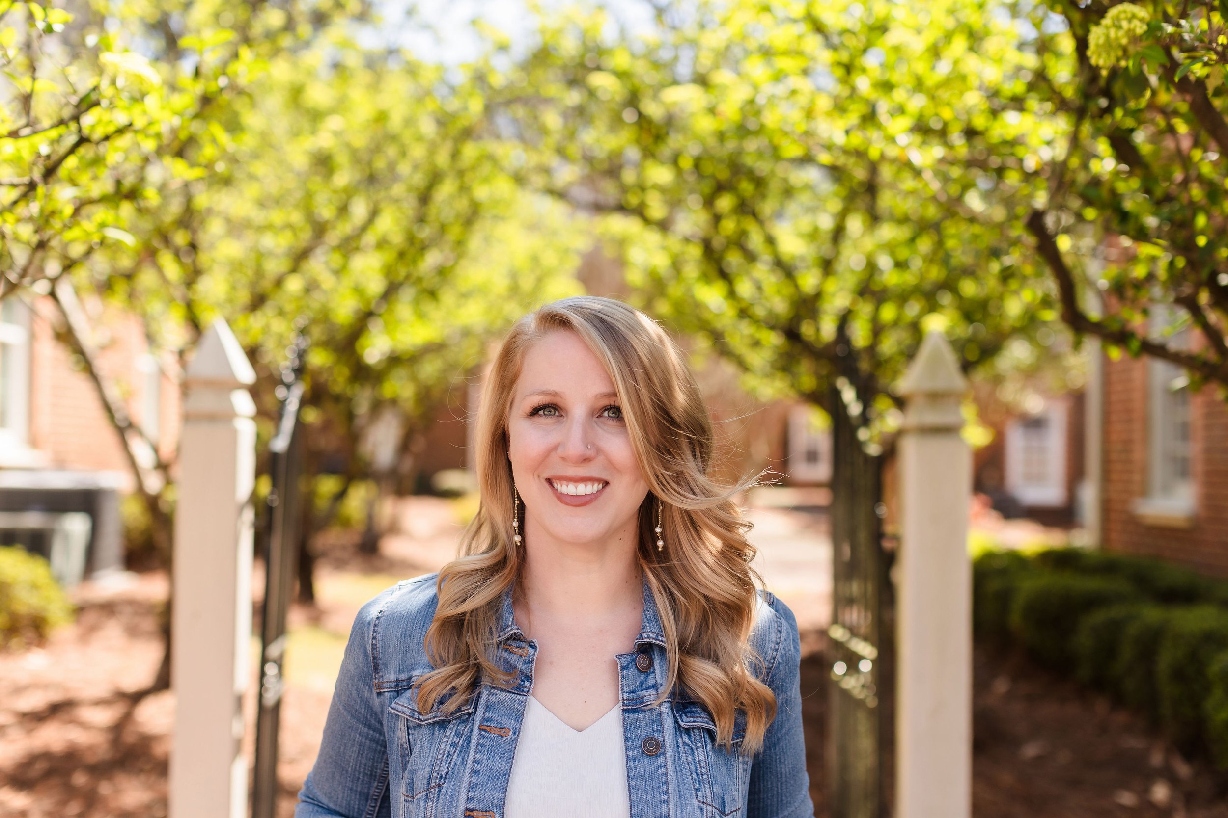 Dr. Jamie L. Chachere, a woman with wavy blonde hair, blue eyes, and earrings, smiling outdoors during daytime with sunlight filtering through green trees in a neighborhood setting.