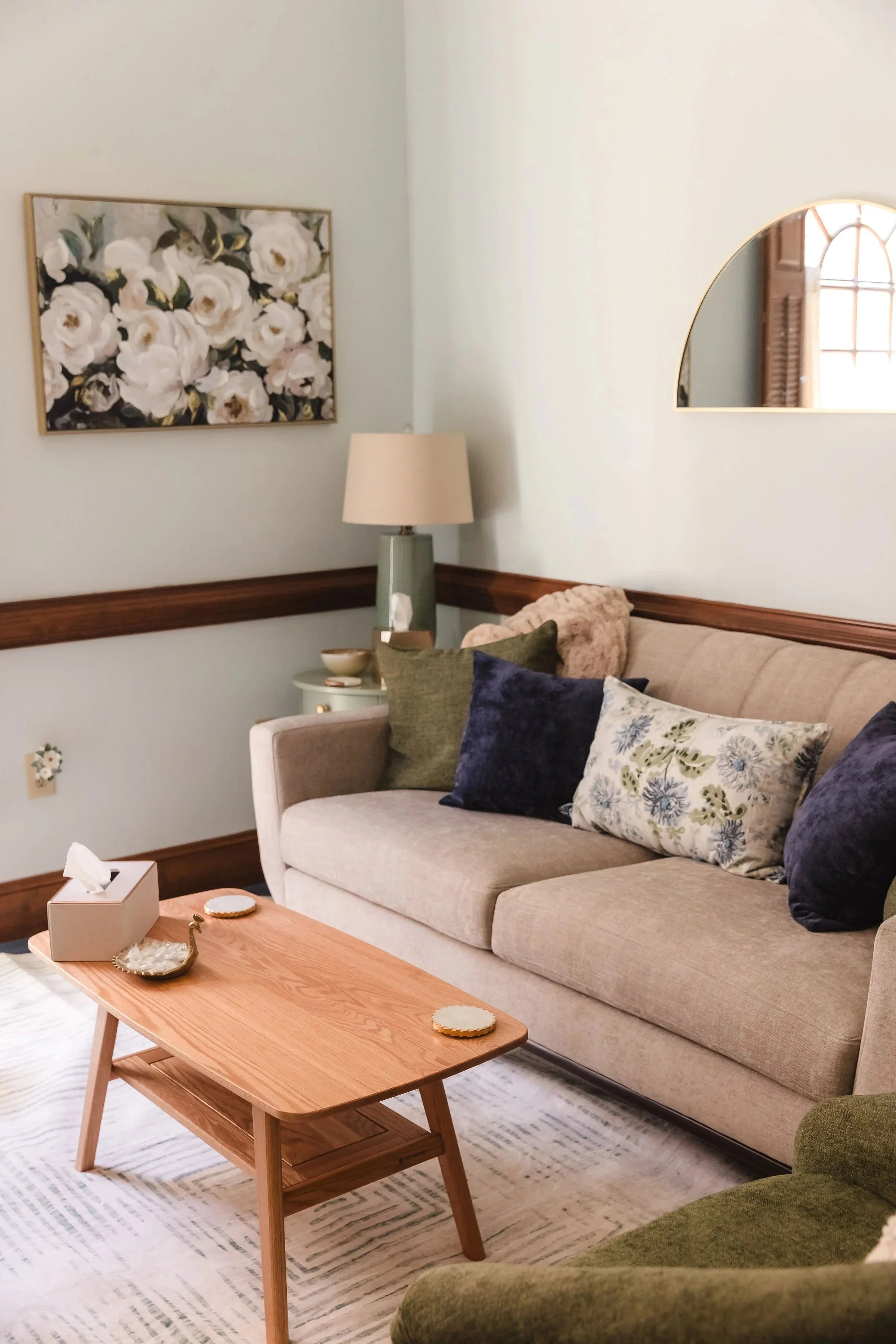 Counseling office with beige sofa and green armchair, a wooden coffee table with coasters, decorative pillows, a lamp, a floral painting on the wall, and a mirror