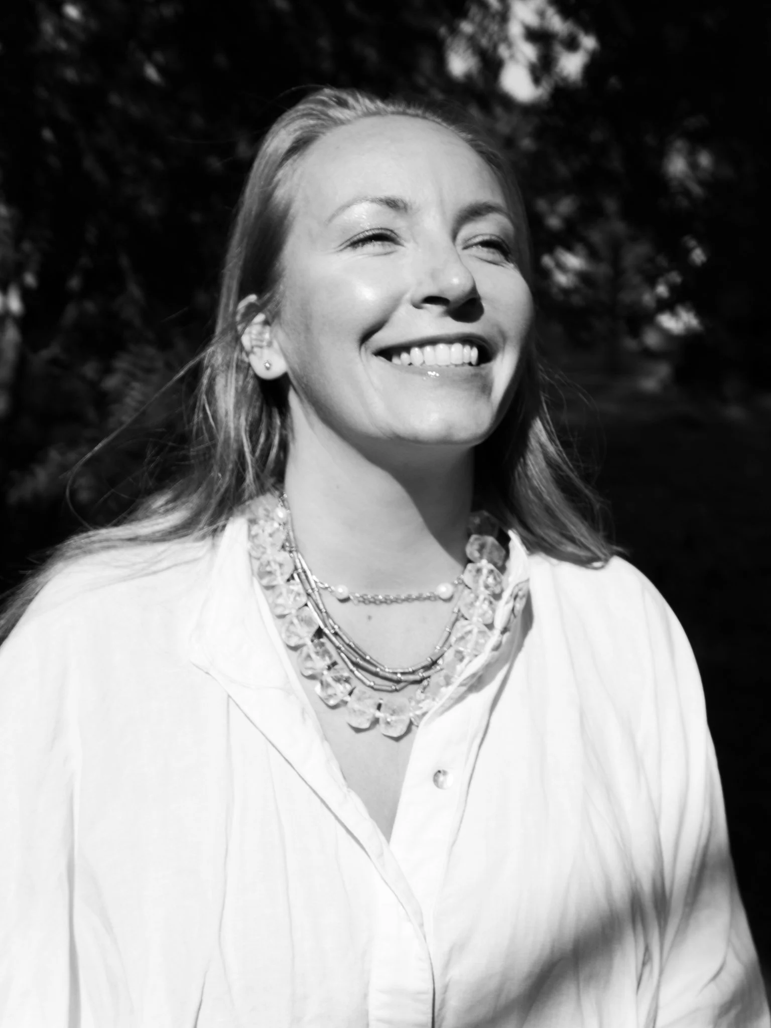 Black and white photo of a smiling woman with light-colored hair, wearing earrings, and a layered necklace, outdoors with blurred trees in the background.
