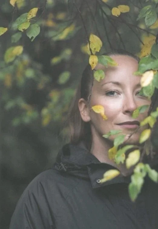 A woman with light skin and brown hair peeking through green and yellow leaves in a forest setting.