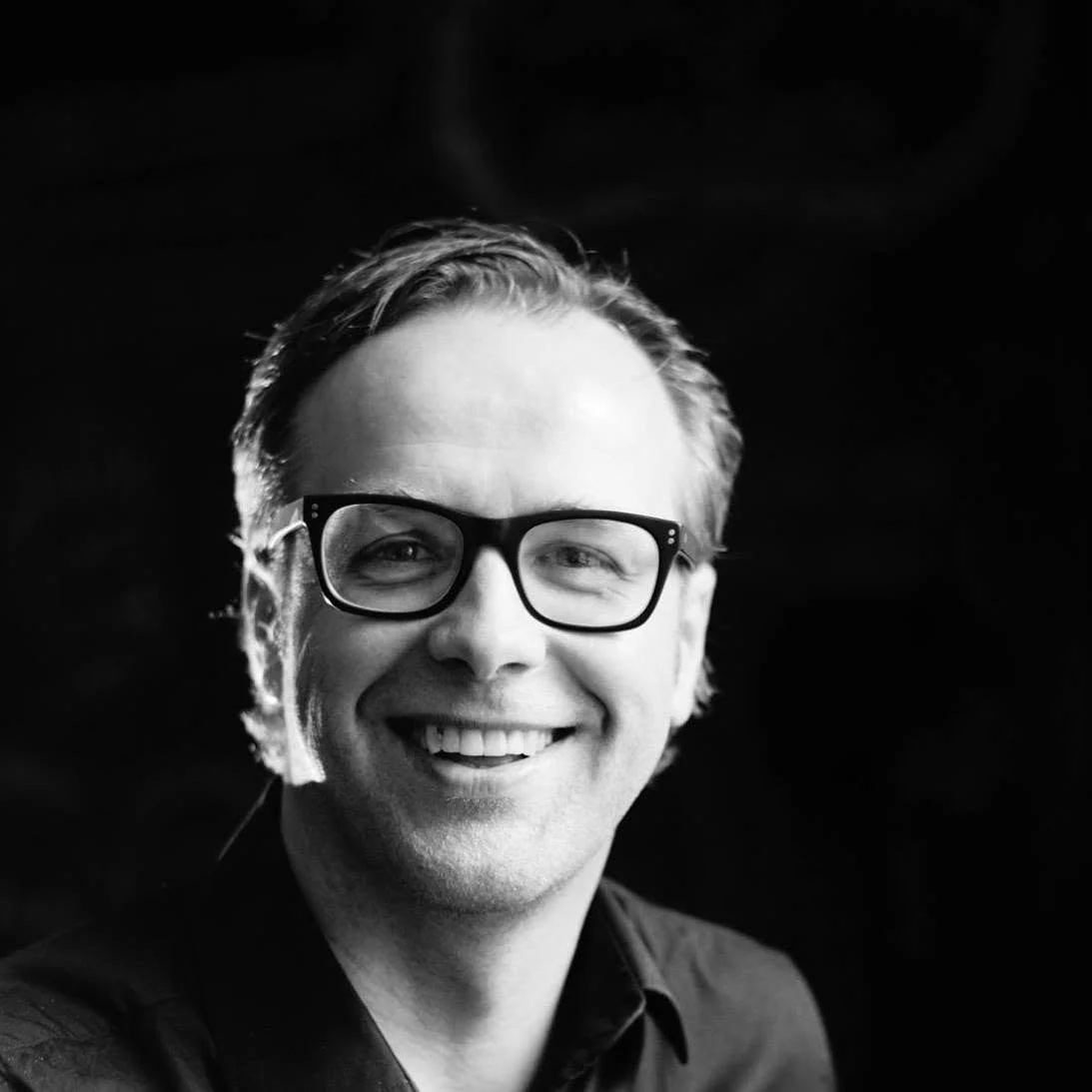 Black and white portrait of a smiling man wearing glasses, dark shirt, and light-colored hair, against a dark background.