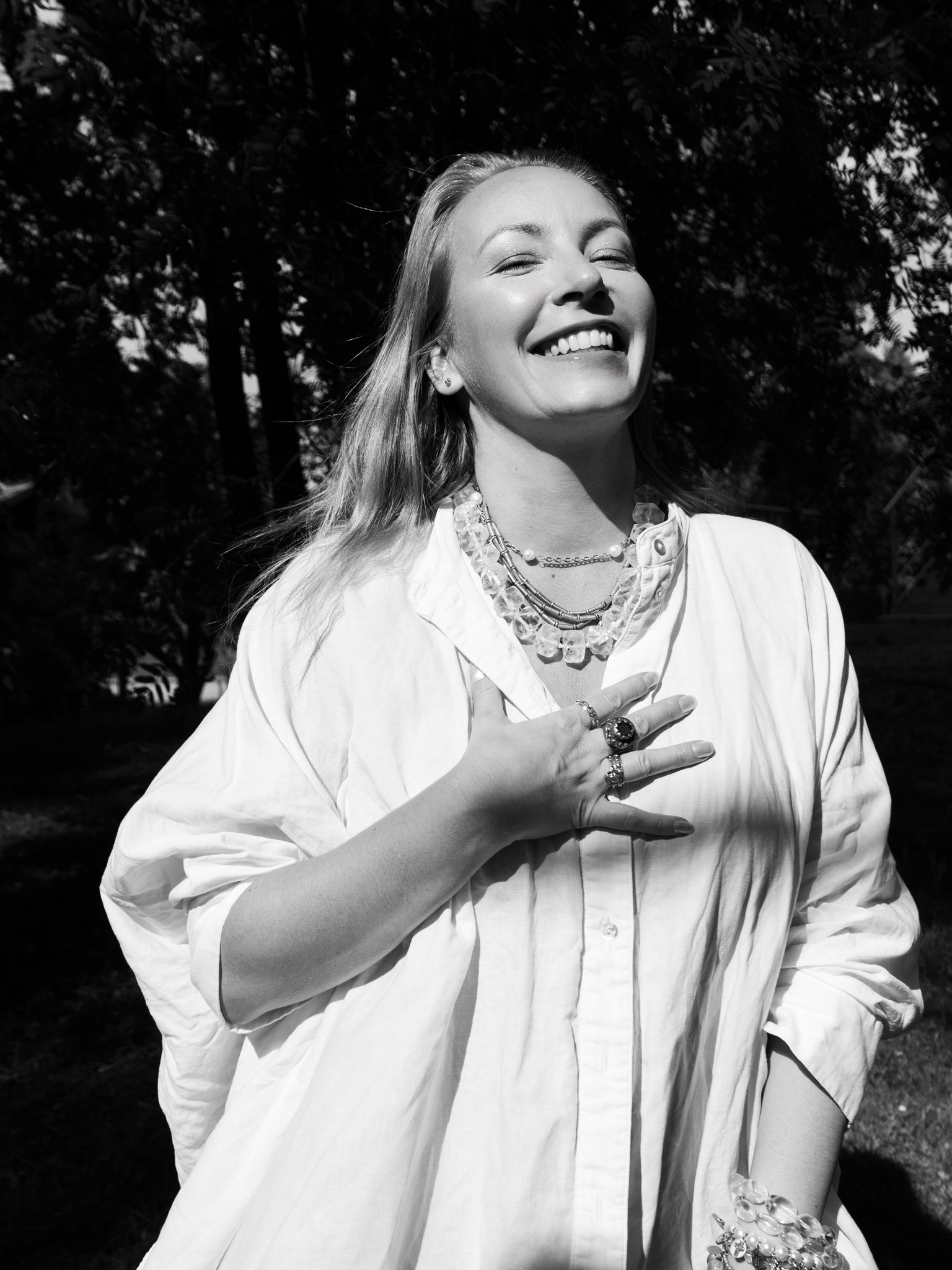 A smiling woman with long hair, wearing a light-colored blouse, multiple necklaces, rings, earrings, and a bracelet, standing outdoors with trees in the background.