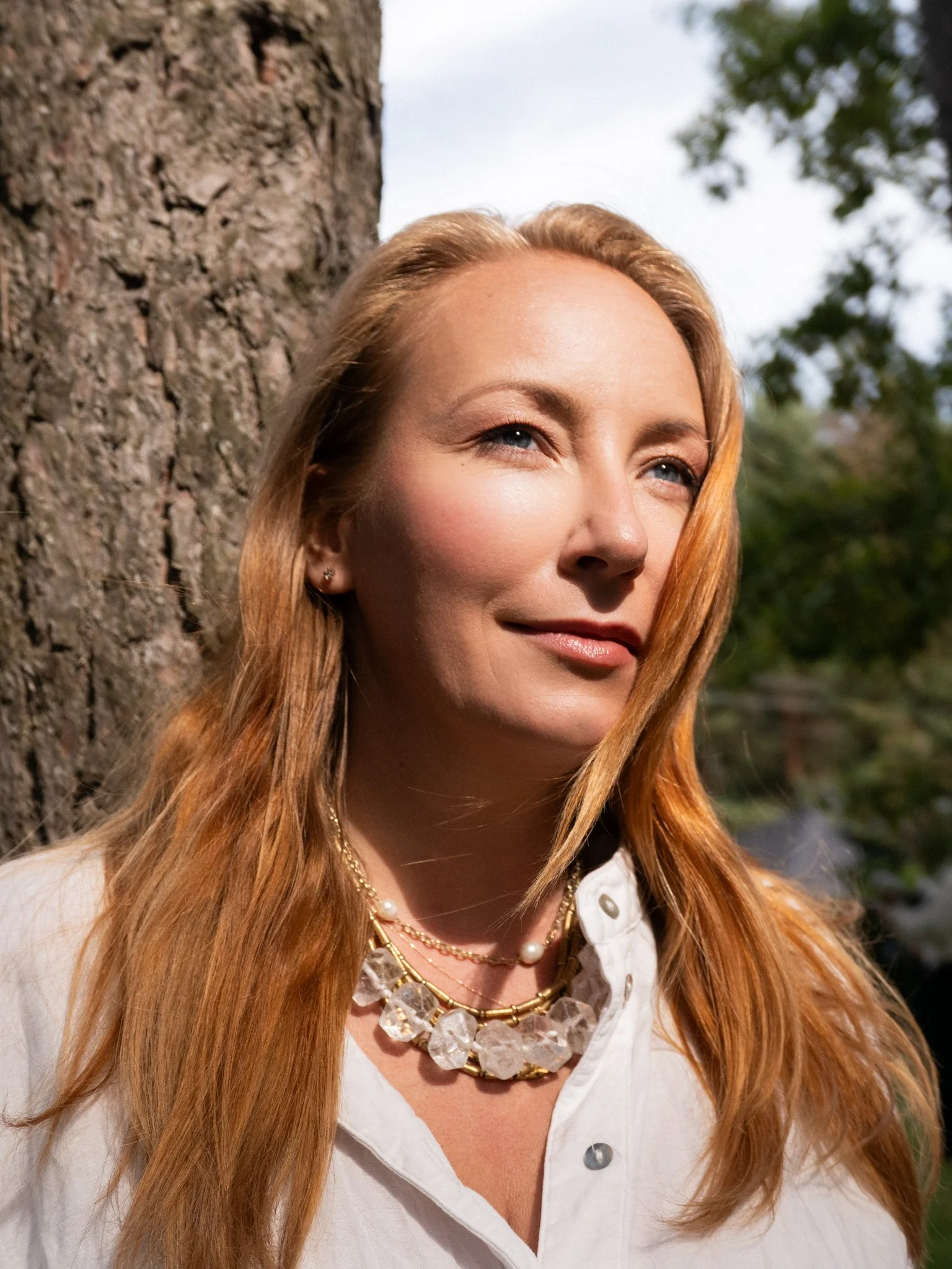 A woman with long red hair standing outdoors near a tree trunk, wearing a white blouse and layered jewelry, looking off to the side with a slight smile.