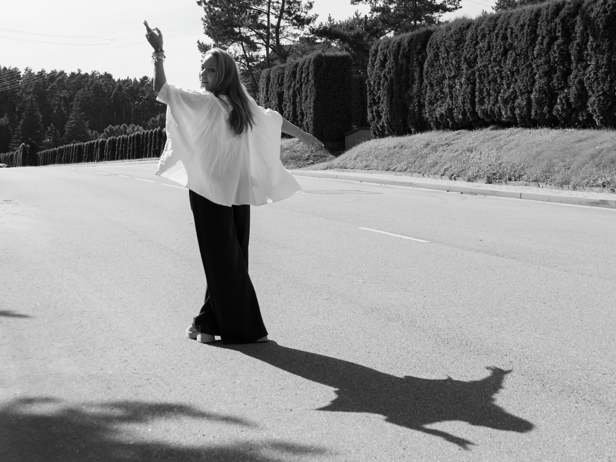 A woman with long hair in a joyful pose on a sunny street with large trees and neatly trimmed bushes in the background, captured in black and white.