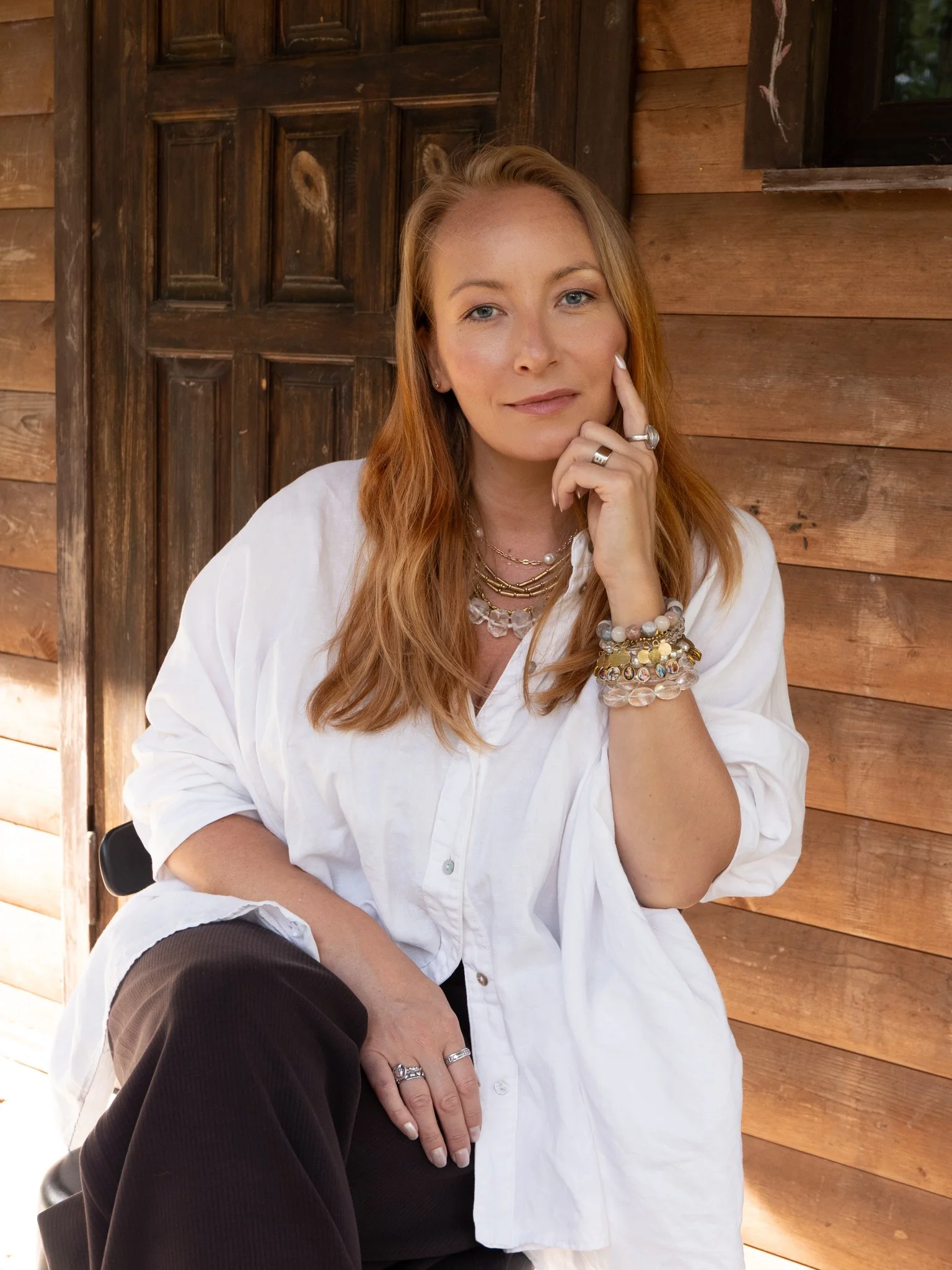 A woman with long reddish-blonde hair sitting in front of a wooden building, touching her face with her right hand and looking at the camera, dressed in a white button-up shirt and wearing various jewelry including rings, necklaces, and bracelets.