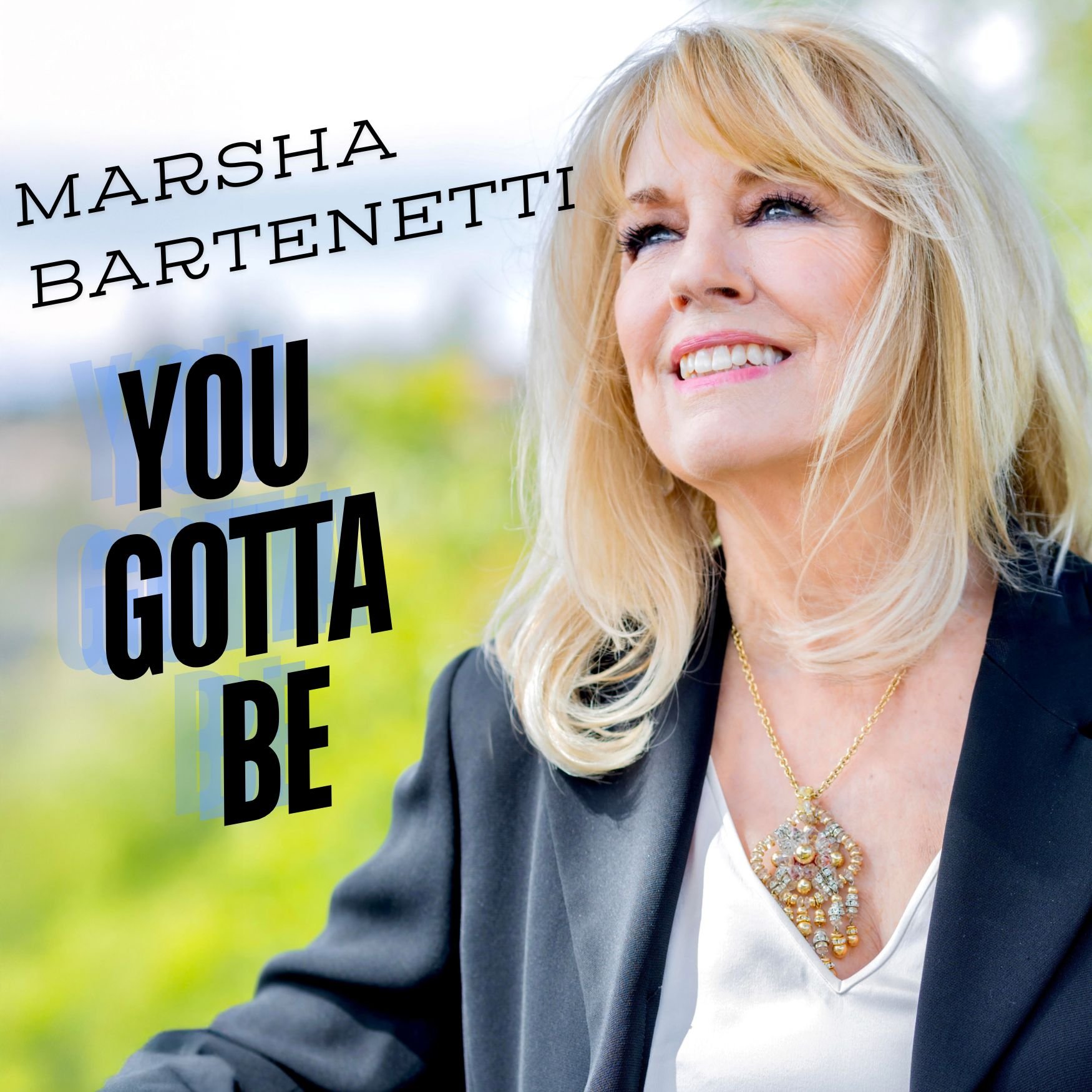 A smiling woman with blonde hair wearing a dark blazer and a gold necklace in an outdoor setting with green trees in the background. Text in the image reads "Marsha Bartenetti" and "You Gotta Be".