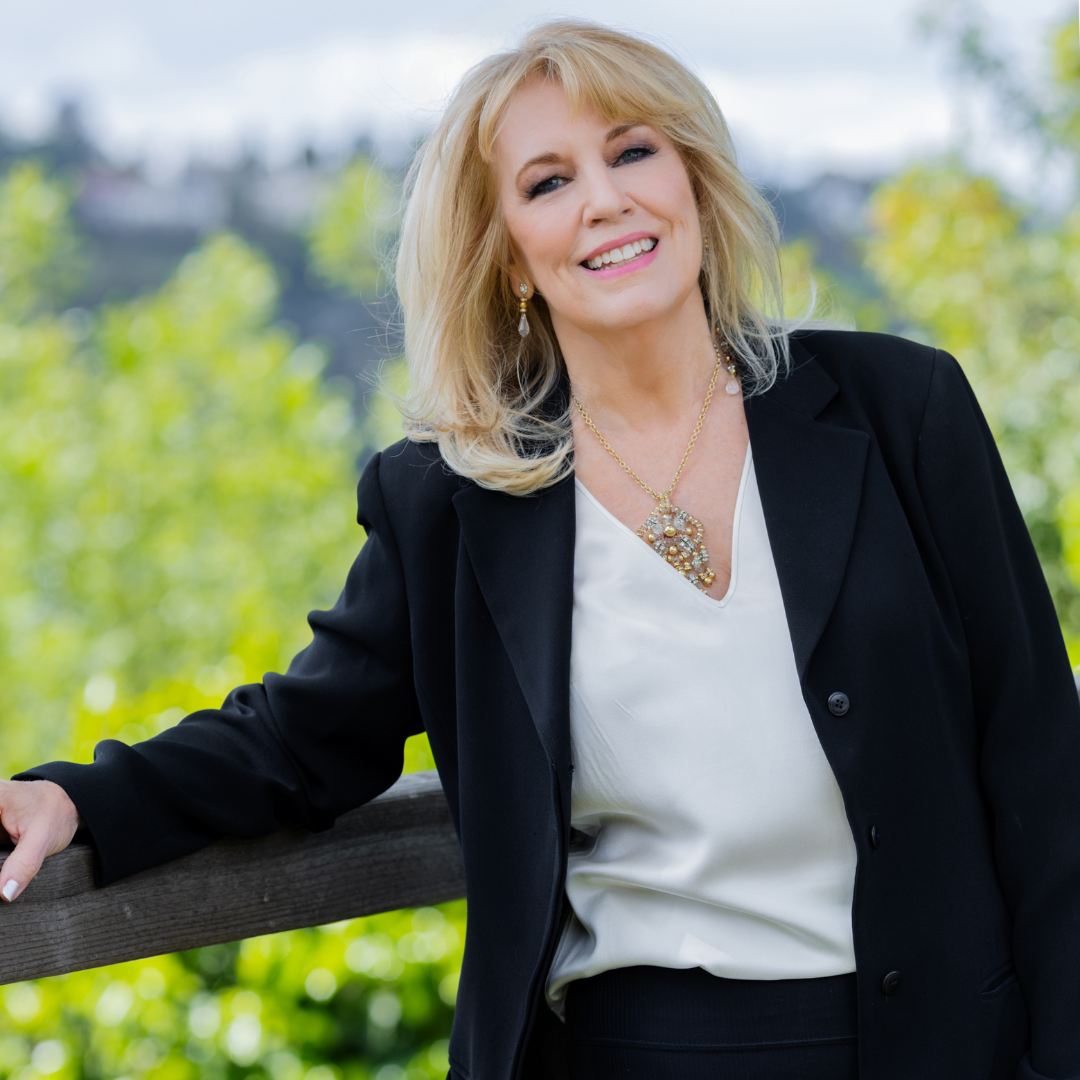 A woman with blonde hair, wearing a black blazer and white blouse, outdoors with green trees in the background, smiling and winking at the camera.