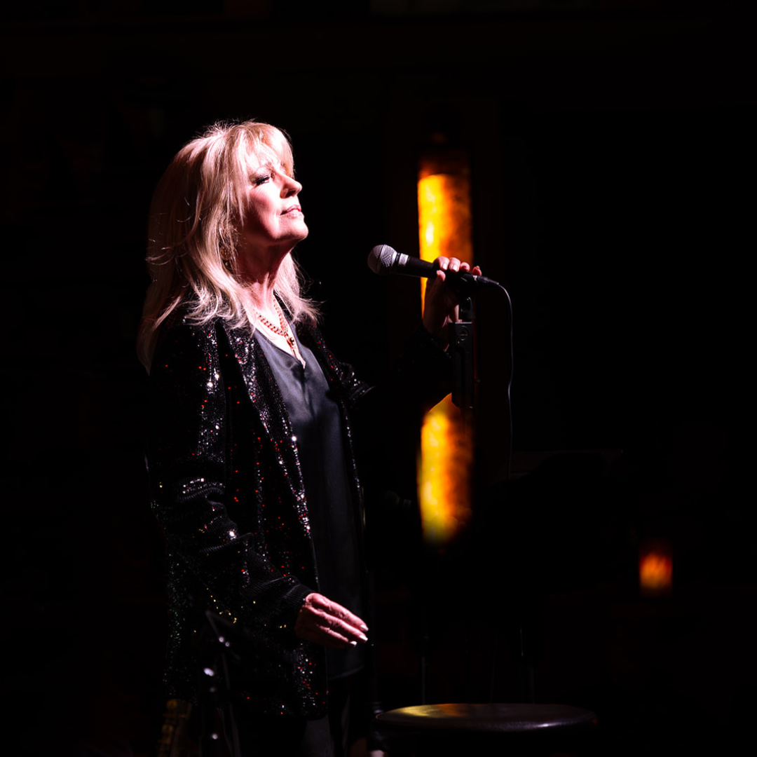 A woman with blonde hair wearing a black, sequin jacket and holding a microphone stands on stage in a dark setting, illuminated by warm light from behind.