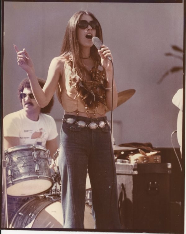 A female singer with long hair, wearing sunglasses, a sleeveless top, and high-waisted jeans, singing into a microphone. A drummer sits behind her, playing a drum set.