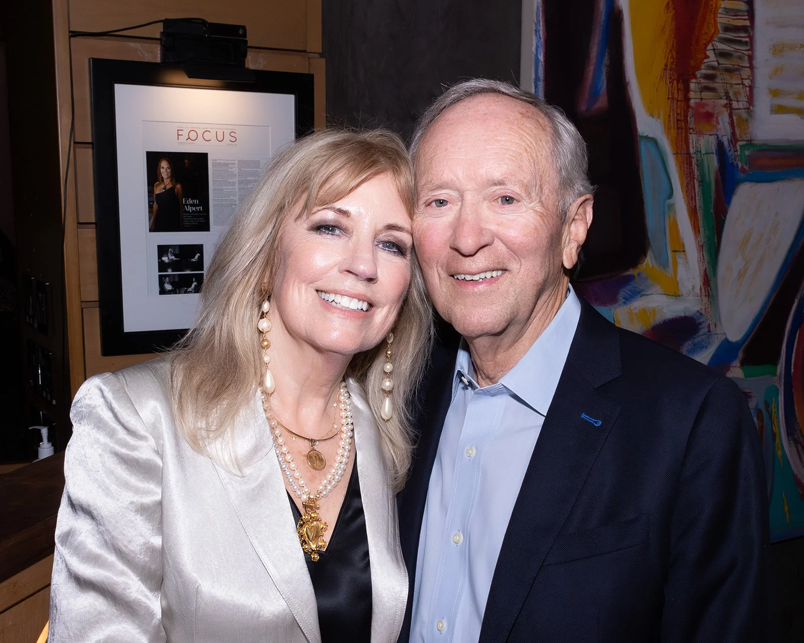 A smiling woman with blonde hair and pearl earrings standing close to an older man with gray hair, both dressed in formal attire, indoors with artwork and a framed magazine article in the background.