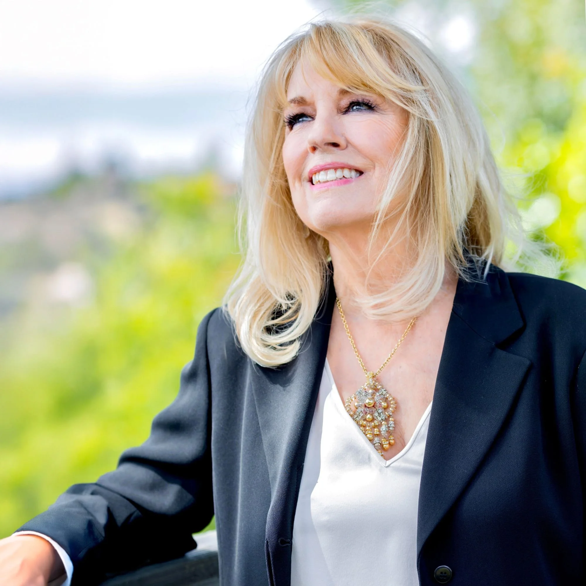 A smiling blonde woman in a black blazer and white top outdoors, with a blurred green background.