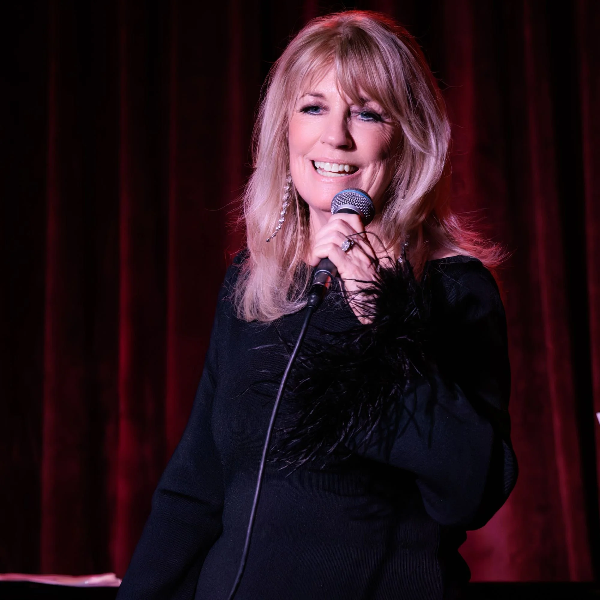 A woman with blonde hair holding a microphone on stage, smiling, in front of a red curtain.