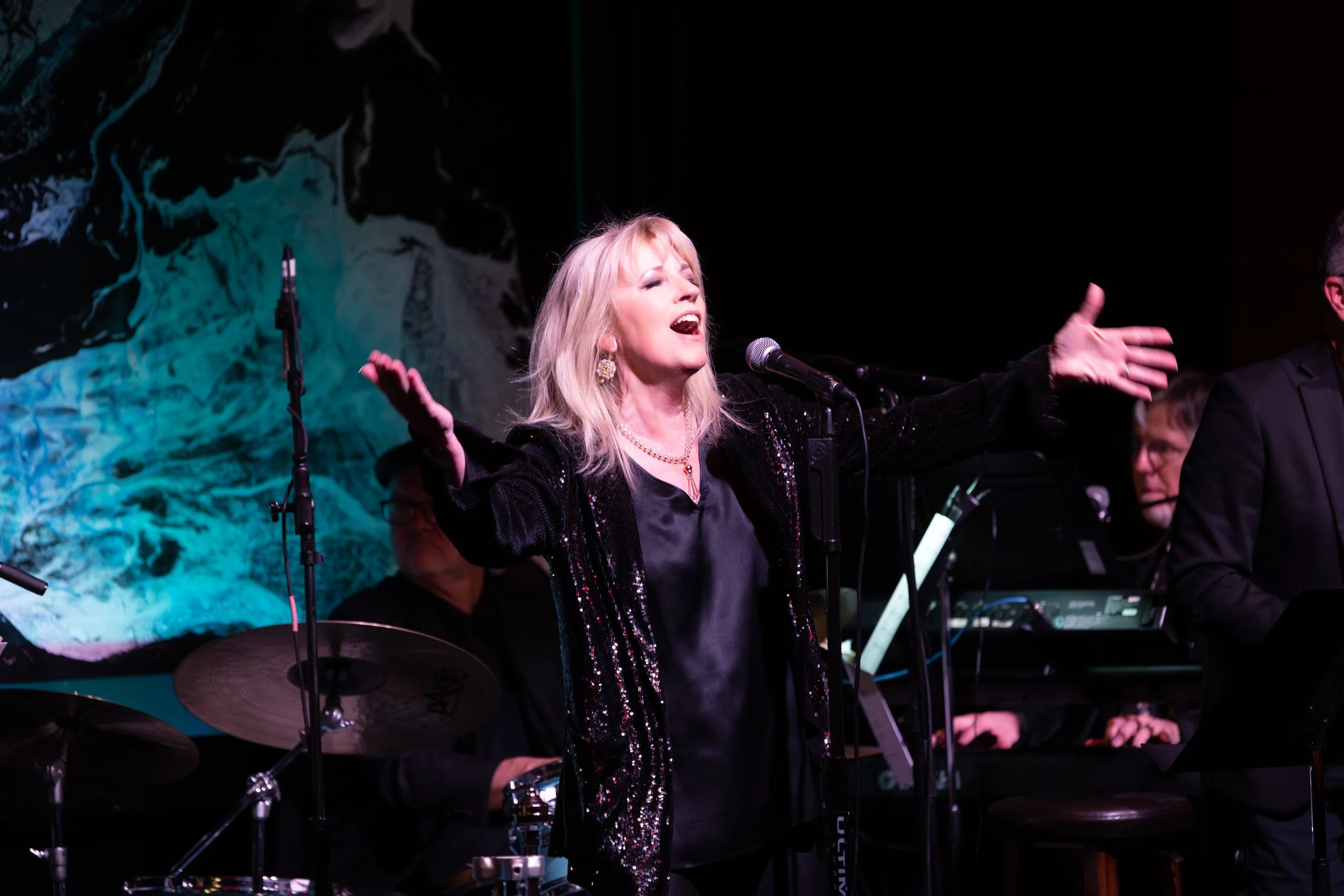 A woman with blonde hair singing passionately on stage with her arms outstretched, accompanied by musicians and a backdrop of an abstract black and blue image.