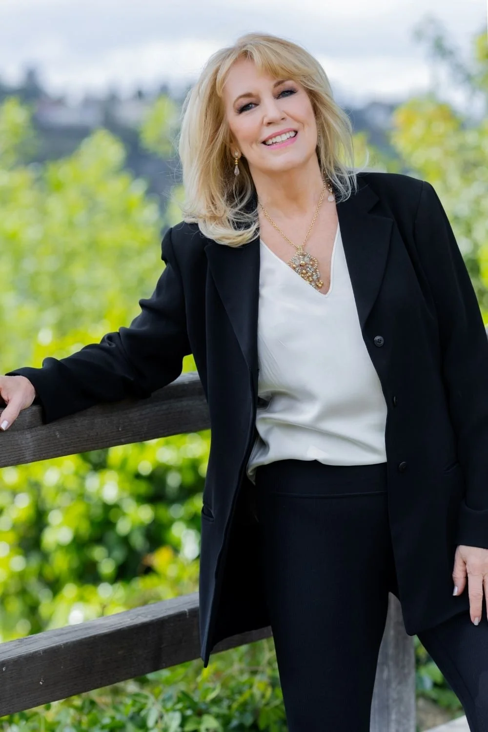A smiling middle-aged woman with blonde hair, wearing a black blazer over a white blouse, and black pants, standing outdoors near a wooden railing with green trees and a cityscape in the background.