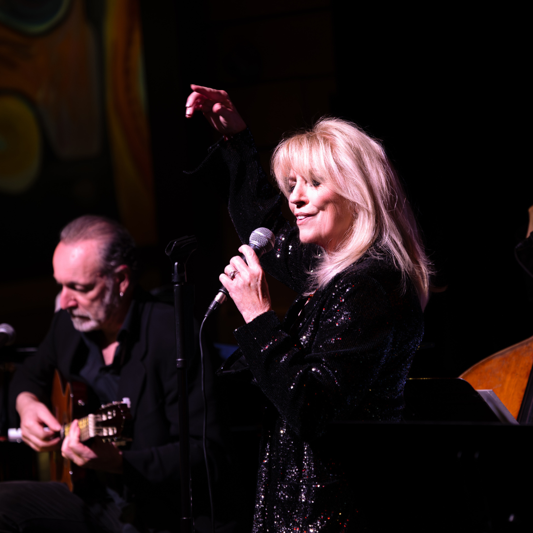 A woman singing into a microphone on stage, accompanied by a man playing a guitar. The woman has blonde hair and is wearing a black, sequined top. The background is dark with some colorful artwork visible.