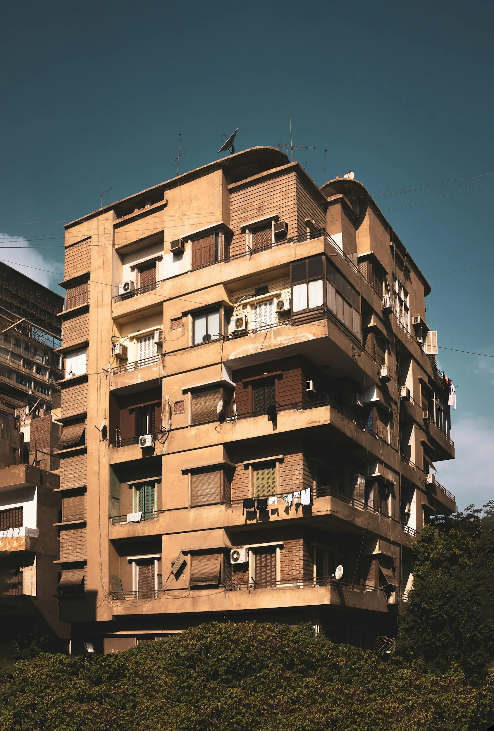 An old, beige apartment building with multiple balconies and air conditioning units, surrounded by clear skies and greenery at the bottom.
