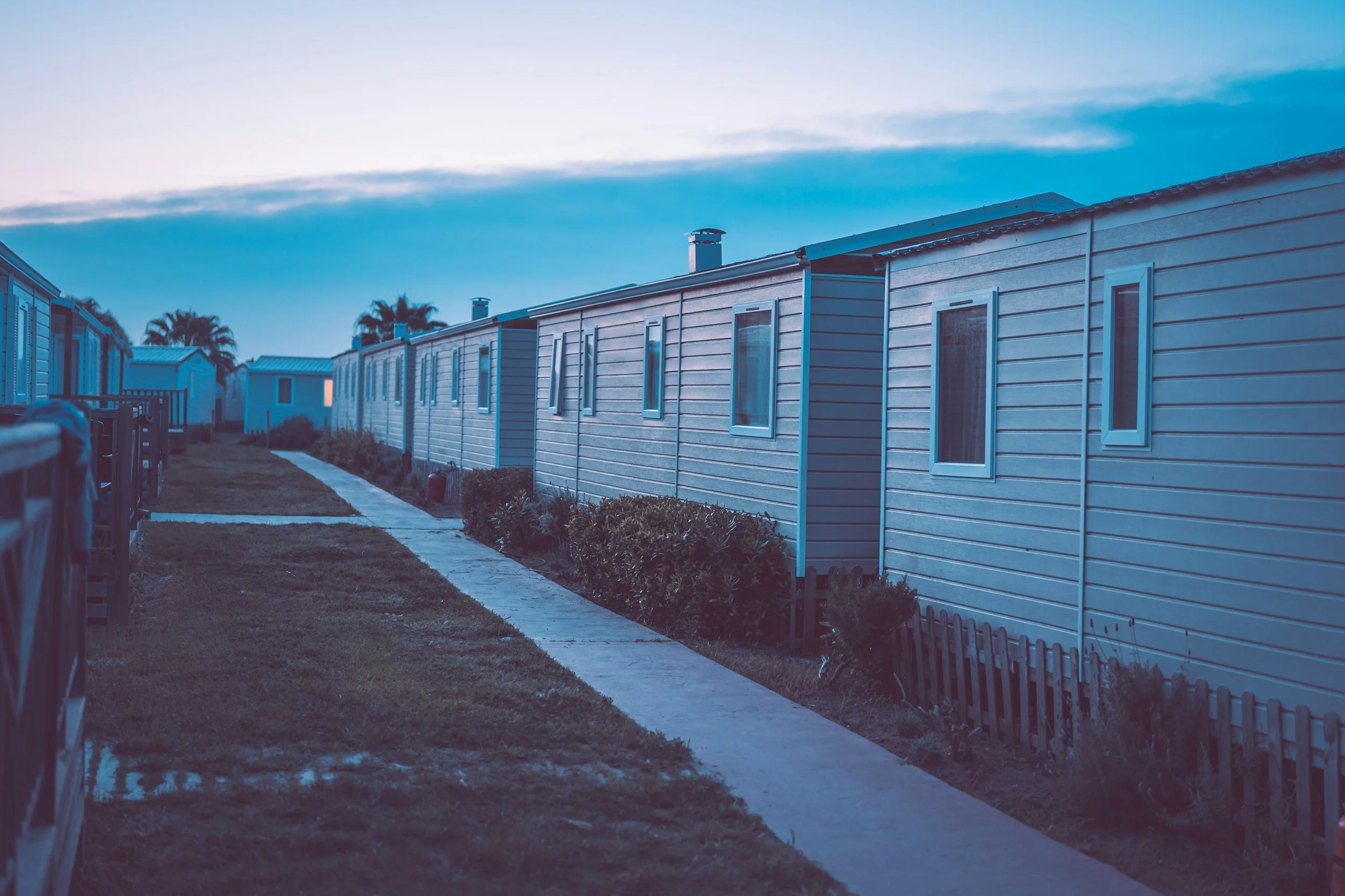 Line of modern mobile homes or trailers with small windows, set along a sidewalk in a residential or park area during dusk or dawn, with palm trees in the background.