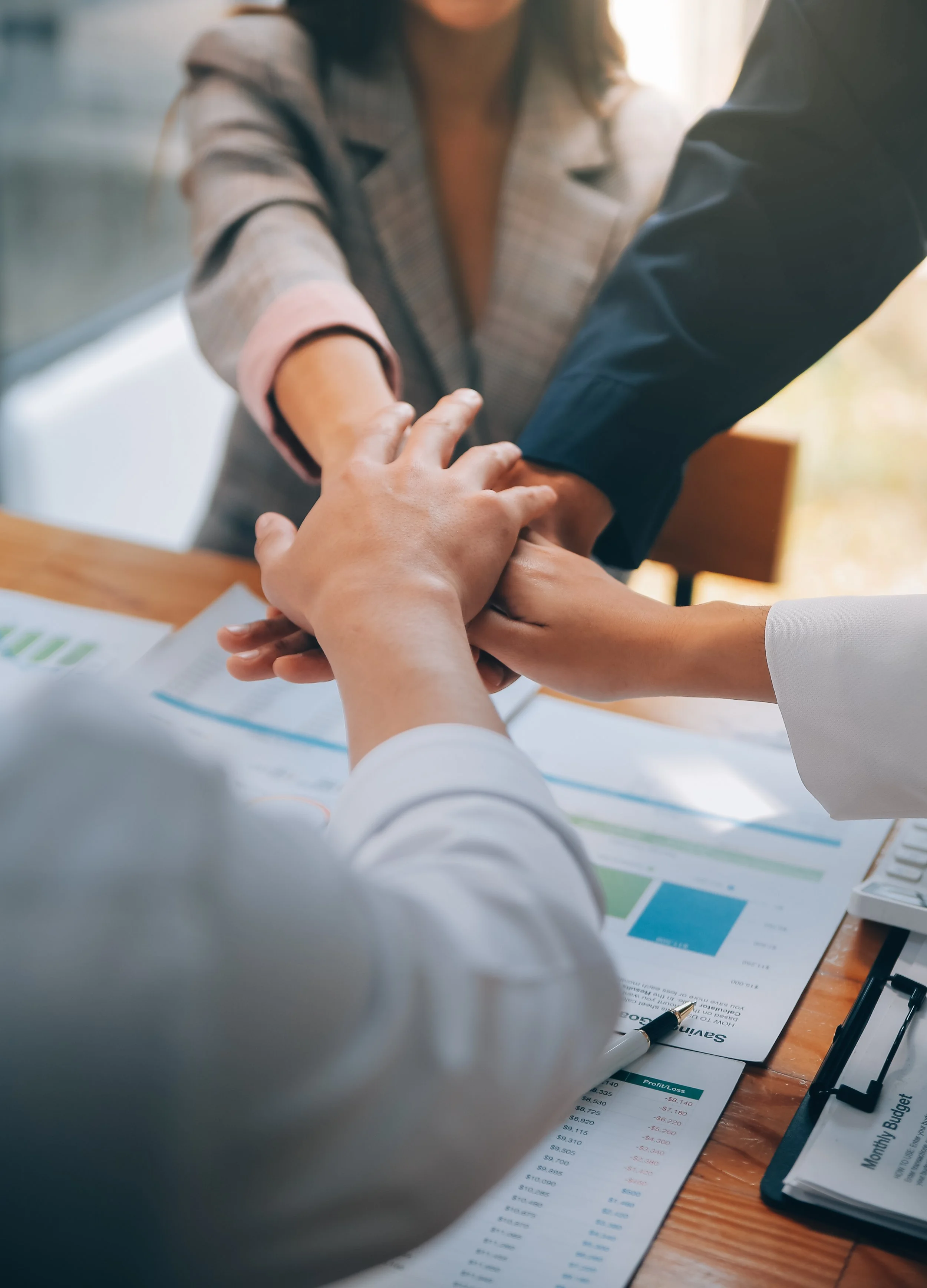 People putting their hands together in a team gesture over a desk with financial documents and a pen.