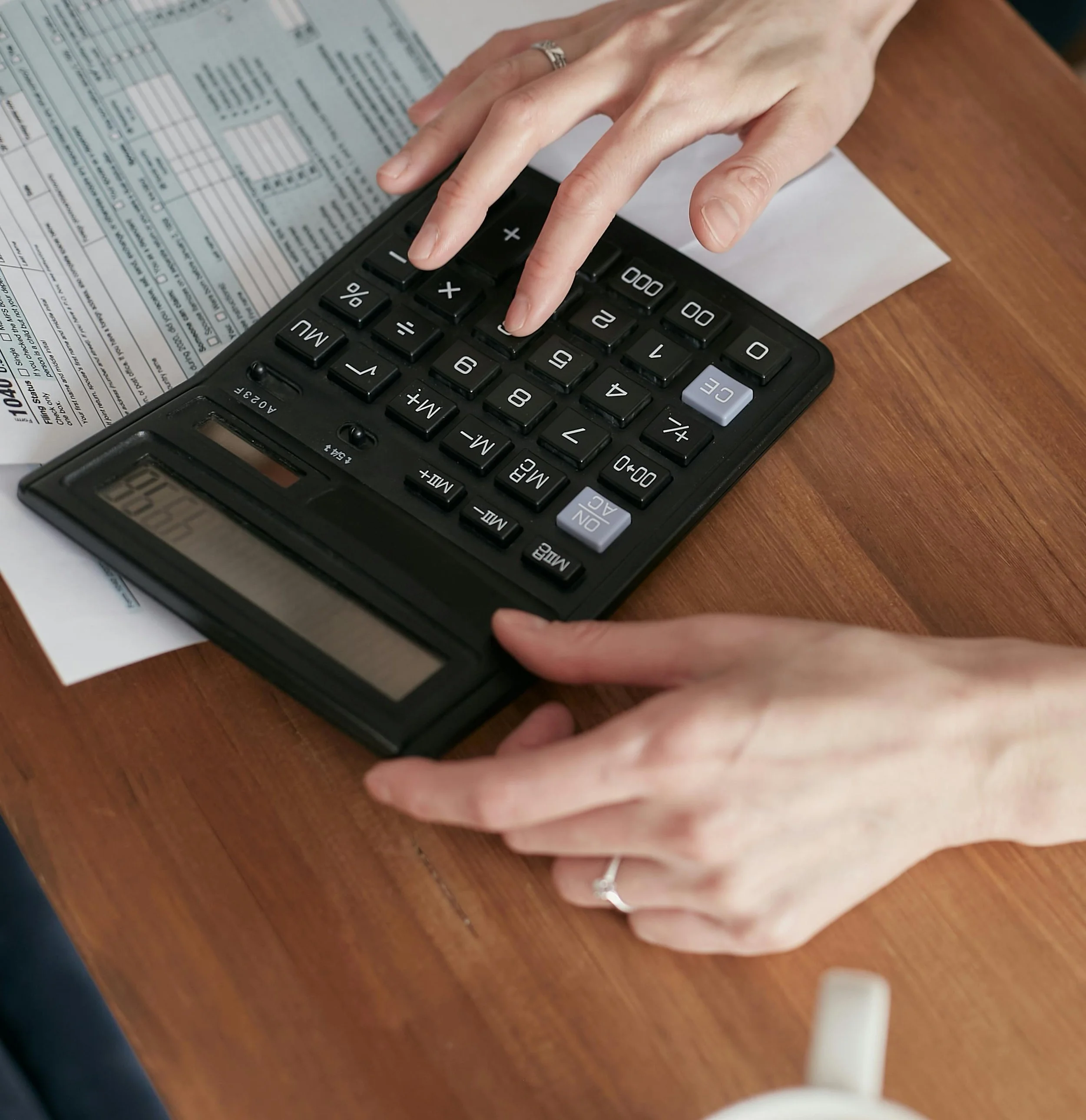 Close-up of a person's hands using a black calculator and a receipt or document on a wooden table.