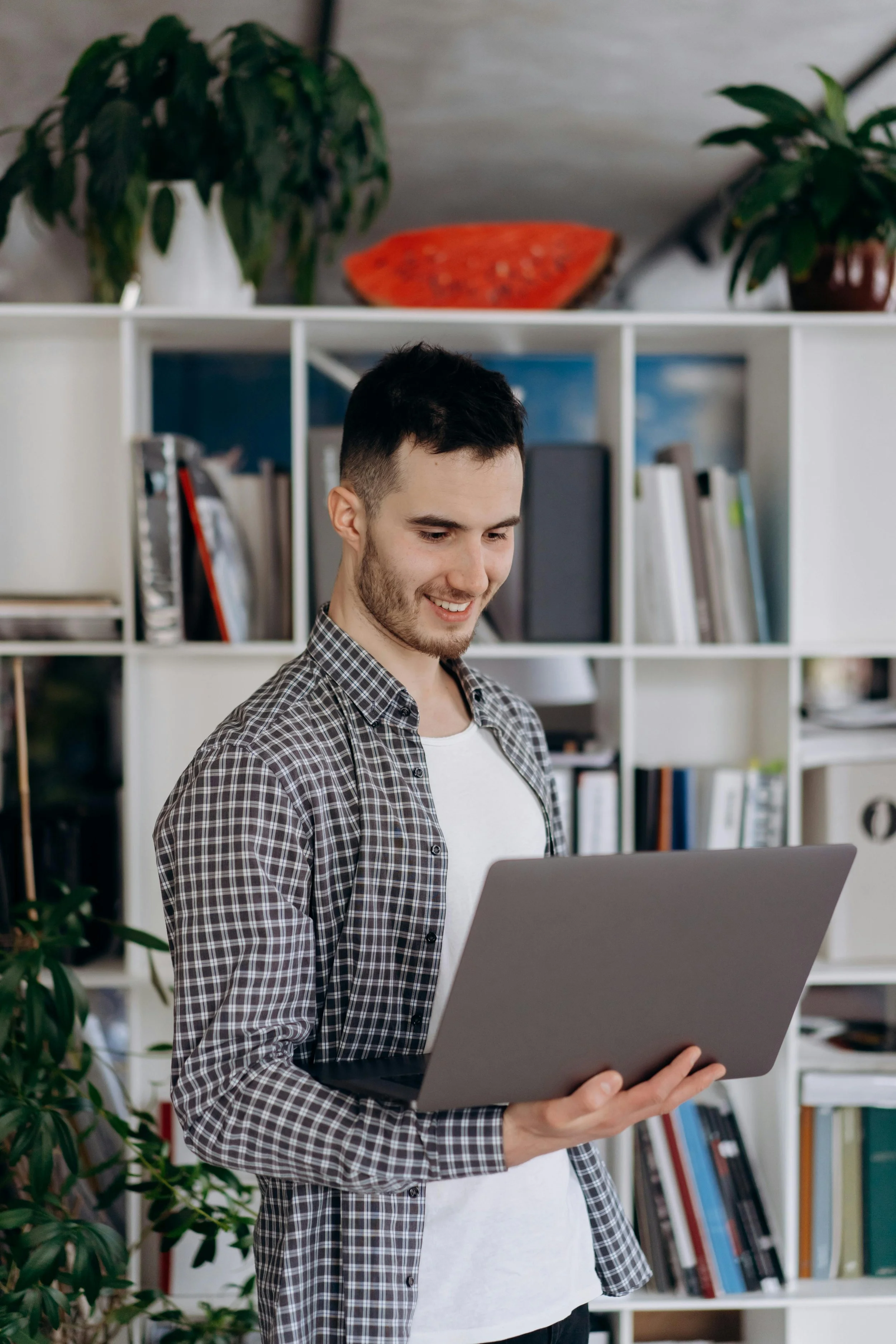 A young man with short dark hair and a beard, wearing a black and white checkered shirt over a white T-shirt, is smiling and looking at a laptop in an indoor setting with a bookshelf and plants in the background.