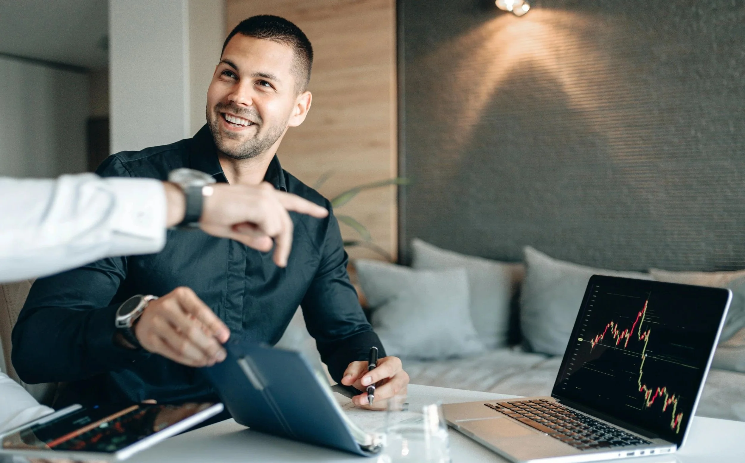 A man with a short beard and dark hair smiling while sitting at a table during a business meeting, with a laptop showing stock market charts, a tablet, and a smartphone nearby.