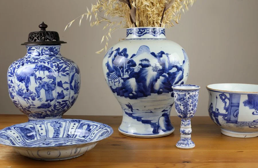 Collection of blue and white porcelain vases, bowls, and glasses on a wooden surface, with a large vase holding dried grass or plants in the background.