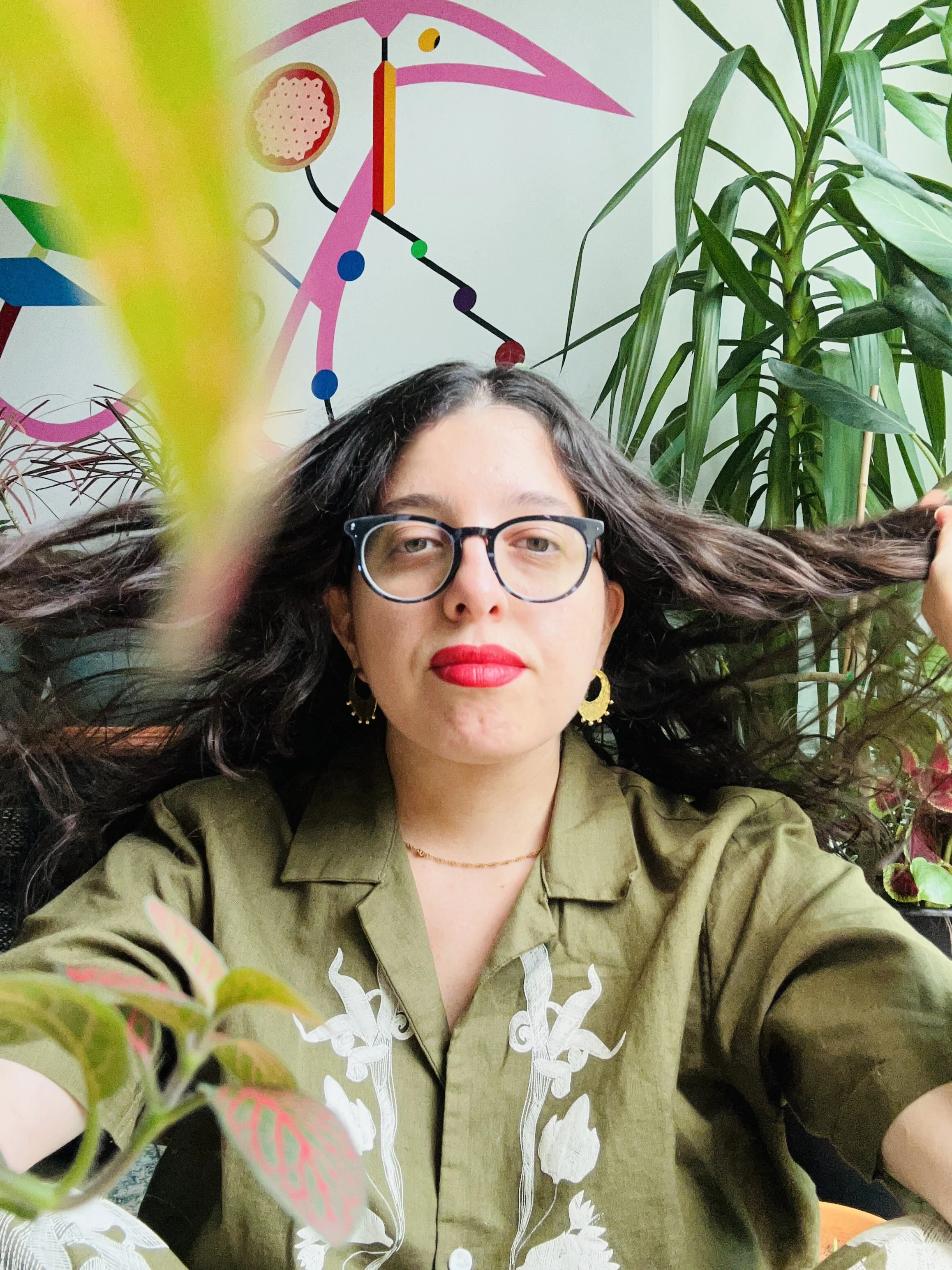 A woman with dark hair, glasses, and red lipstick, wearing a green shirt with white floral embroidery, sitting in front of a colorful abstract wall with plants and a stylized pink bird design.