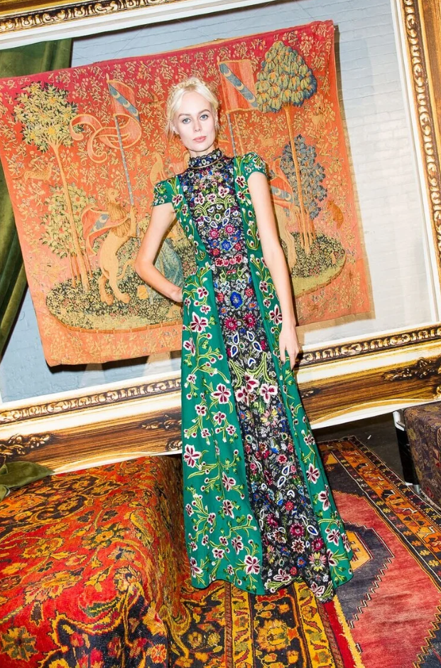 A woman standing in front of a detailed tapestry with trees and animals, dressed in a long, colorful floral-patterned dress with a green outer layer, in a room with ornate furniture and a patterned rug.