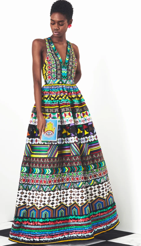 Woman in a colorful, patterned, sleeveless maxi dress with intricate embroidery, standing against a plain white background, holding a small clutch with one hand.