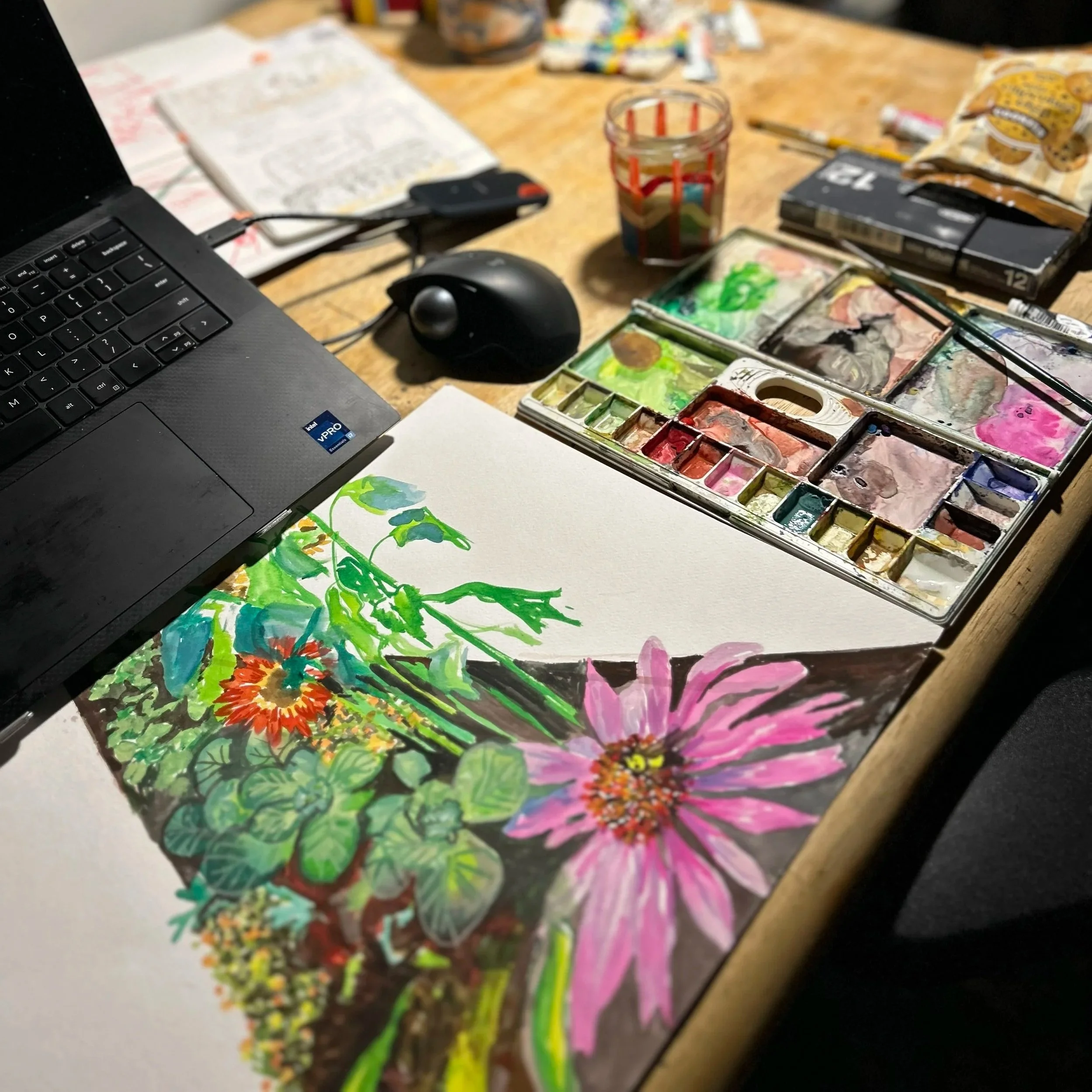 A watercolor painting of pink and orange flowers with green leaves on white paper, surrounded by paint supplies, a laptop, and art tools on a wooden table.