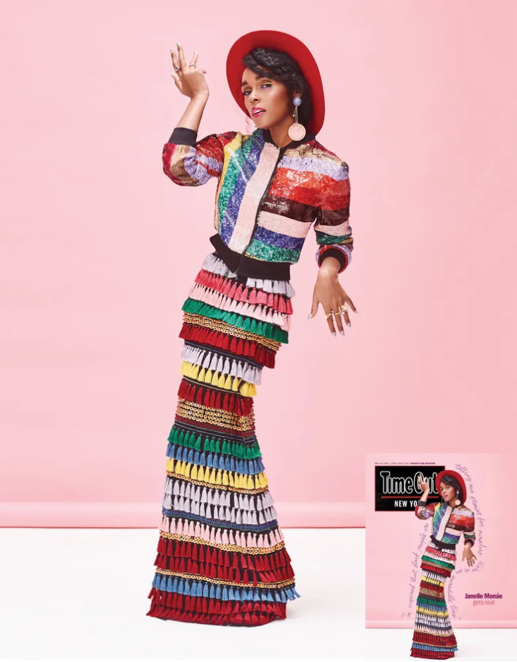 A woman in a colorful, patterned outfit with a rainbow-colored skirt, a multicolored top, a red hat, and large earrings, standing against a pink background.