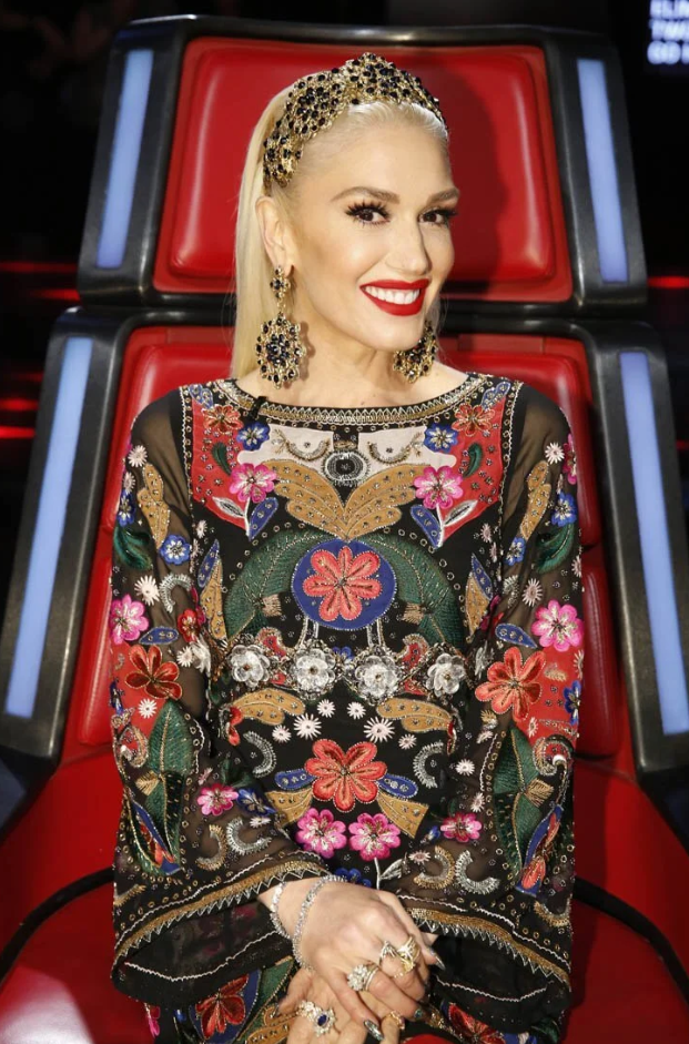 Gwen Stefani a singer with blonde hair and red lipstick sitting on a red chair, wearing a colorful embroidered black dress with floral and intricate patterns, large earrings, and a gold headband with black embellishments.