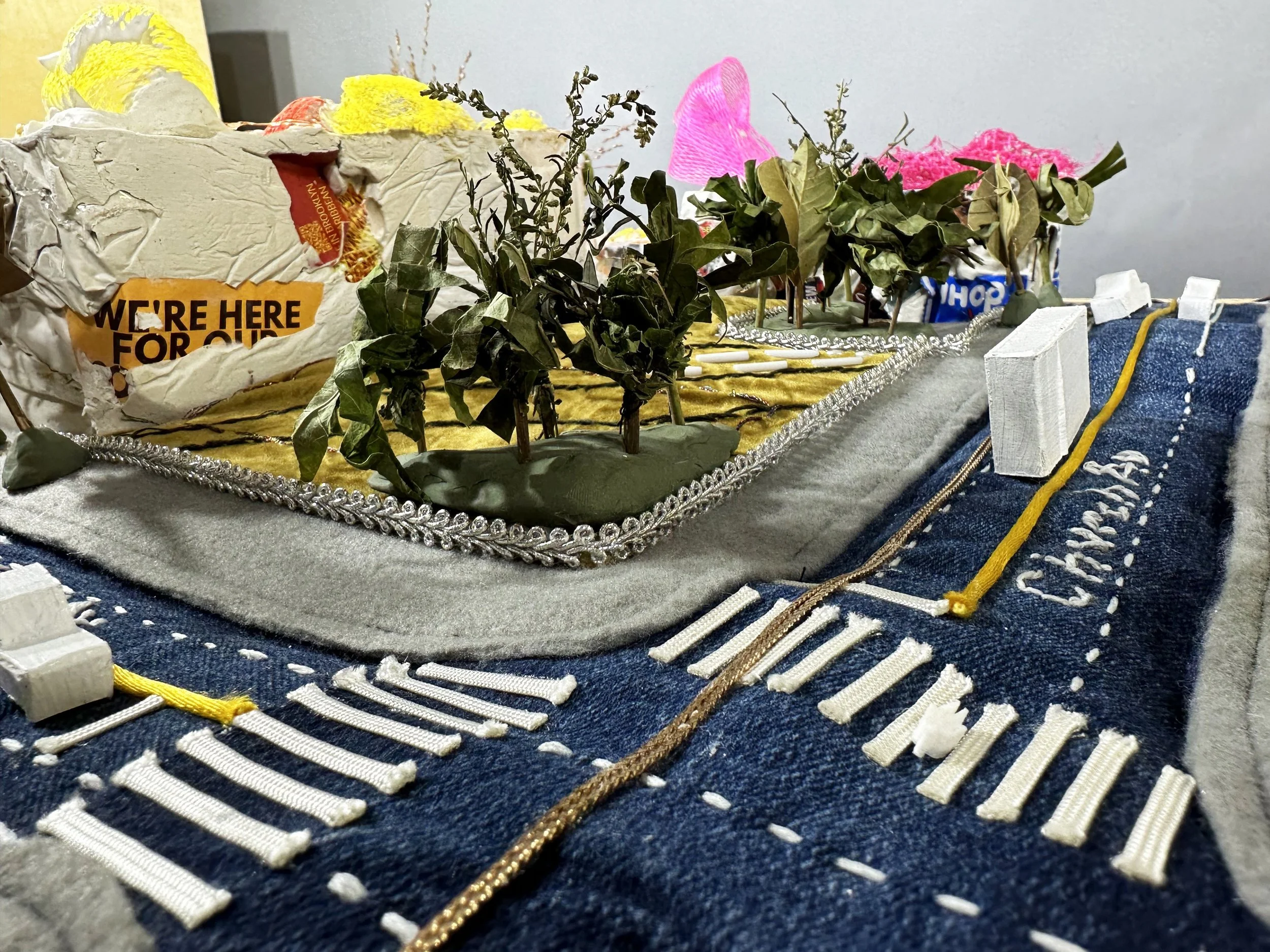 A detailed diorama of a city street made from textiles and craft materials, featuring trees, a road with traffic lines, small buildings, and various decorative elements.