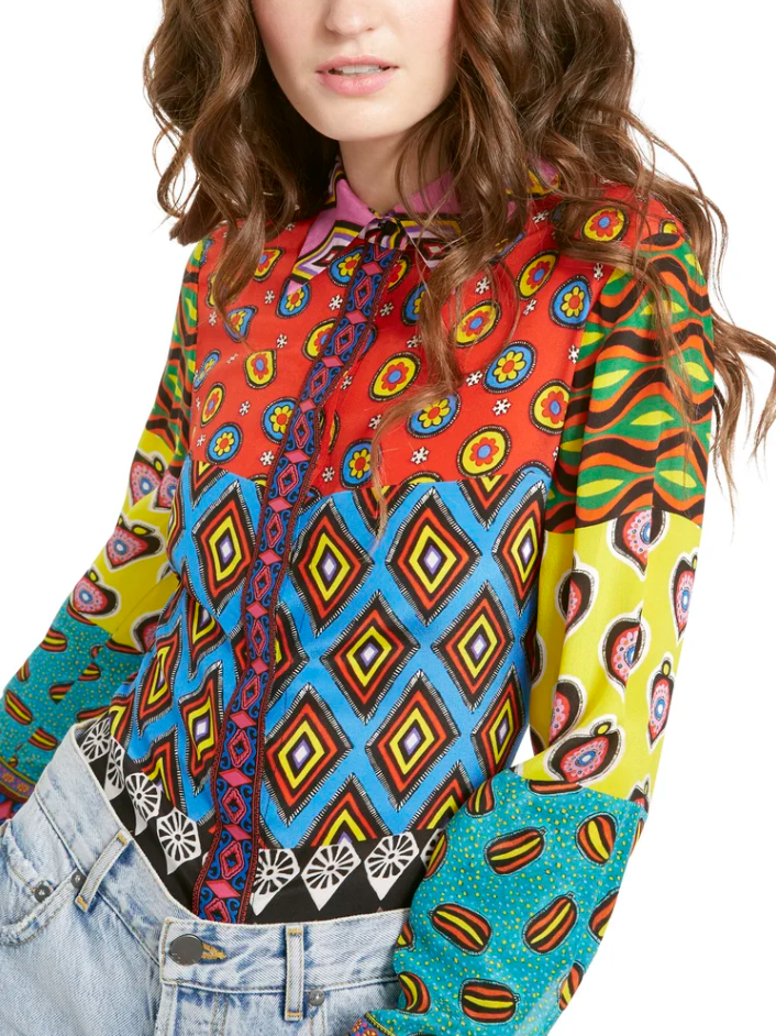 Woman wearing a colorful, patterned blouse with floral, geometric, and abstract designs in red, blue, yellow, green, and black, paired with light blue jeans.